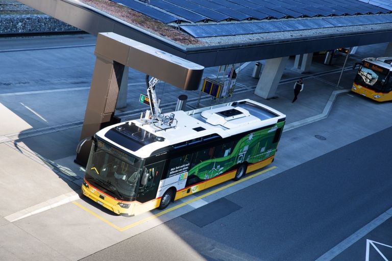 Luftaufnahme eines Elektrobusses an einer Ladestation. Der Bus hat ein weißes Dach, eine schwarze Front und grüne und gelbe Farben mit Grafiken auf der Karosserie. Ein zweiter Bus ist weiter entfernt unter einem überdachten Bereich geparkt. Eine Person geht in der Nähe der Station.