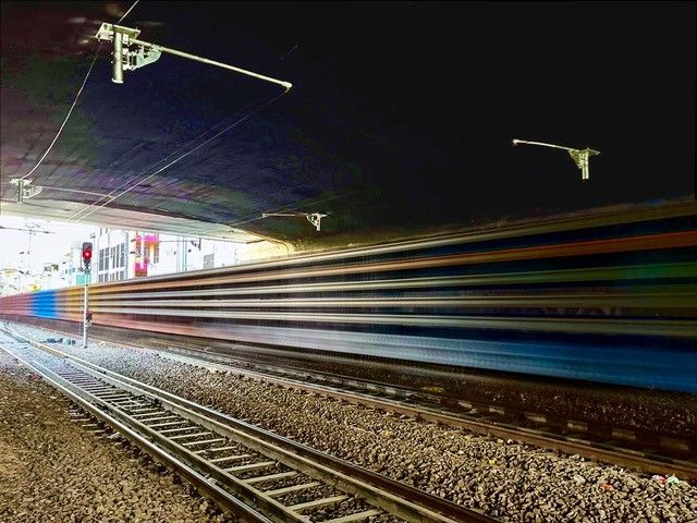 Unscharfe Langzeitbelichtung eines bunten Zuges, der unter einer dunklen Brücke hindurchfährt. Die Bewegung des Zuges erzeugt horizontale Lichtstreifen. Die Gleise und ein rotes Signallicht auf der linken Seite sind sichtbar. Sonnenlicht fällt durch Öffnungen im Hintergrund ein.