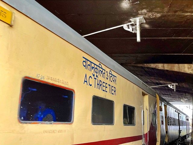 Ein Zugwaggon mit der Aufschrift „AC THREE TIER“ und „वातानुकूलित 3 tier“ auf Hindi steht unter einem Bahnsteig in einem Bahnhof. Der Waggon hat blau getönte Fenster und scheint Teil eines klimatisierten Personenzugs zu sein.  .