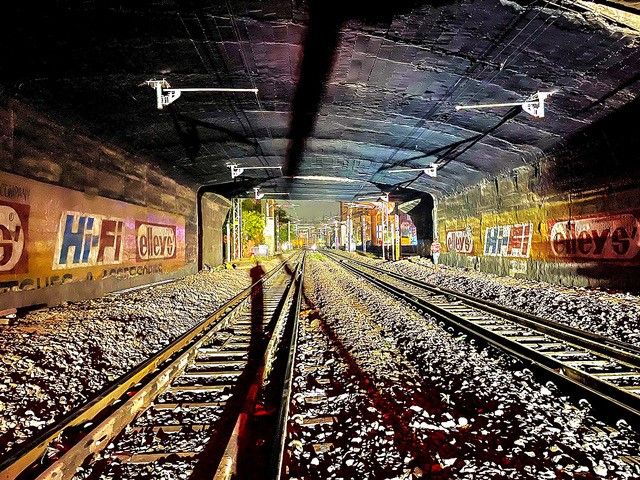Ein schwach beleuchteter unterirdischer Eisenbahntunnel mit zwei parallelen Gleisen, die sich in die Ferne erstrecken. Die Wände sind mit bunten Graffiti geschmückt, und am Ende des Tunnels ist ein schwaches Licht sichtbar, das auf einen Ausgang oder einen weiteren Gleisabschnitt hindeutet.