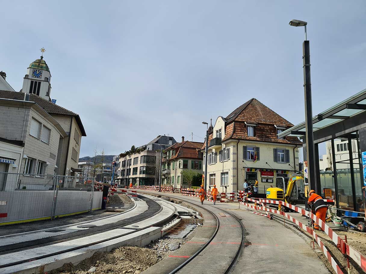 Eine Straße wird gerade gebaut, mehrere Arbeiter in orangefarbener Schutzausrüstung. Die Szene zeigt teilweise fertiggestellte Straßenbahnschienen, Bauzäune und Bauzäune. Gebäude, darunter eine große Kirche und verschiedene Wohn- und Geschäftsgebäude, säumen die Straße.