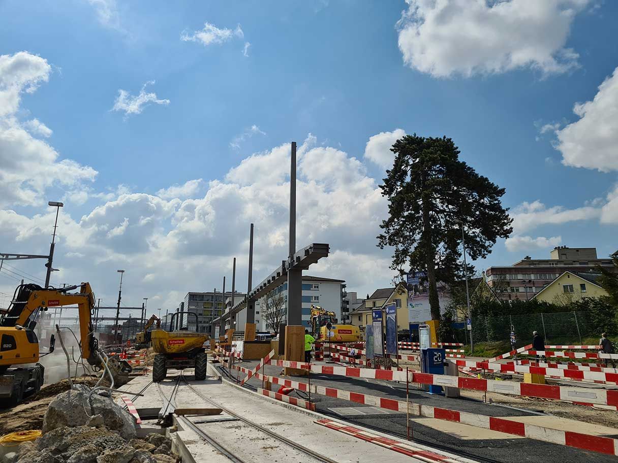 Eine Baustelle mit schweren Maschinen und Geräten. Metallträger werden hochgezogen und der Bereich ist von Sicherheitsbarrieren und Schildern umgeben. Im Hintergrund sind Wohnhäuser und Bäume unter einem teilweise bewölkten Himmel zu sehen.