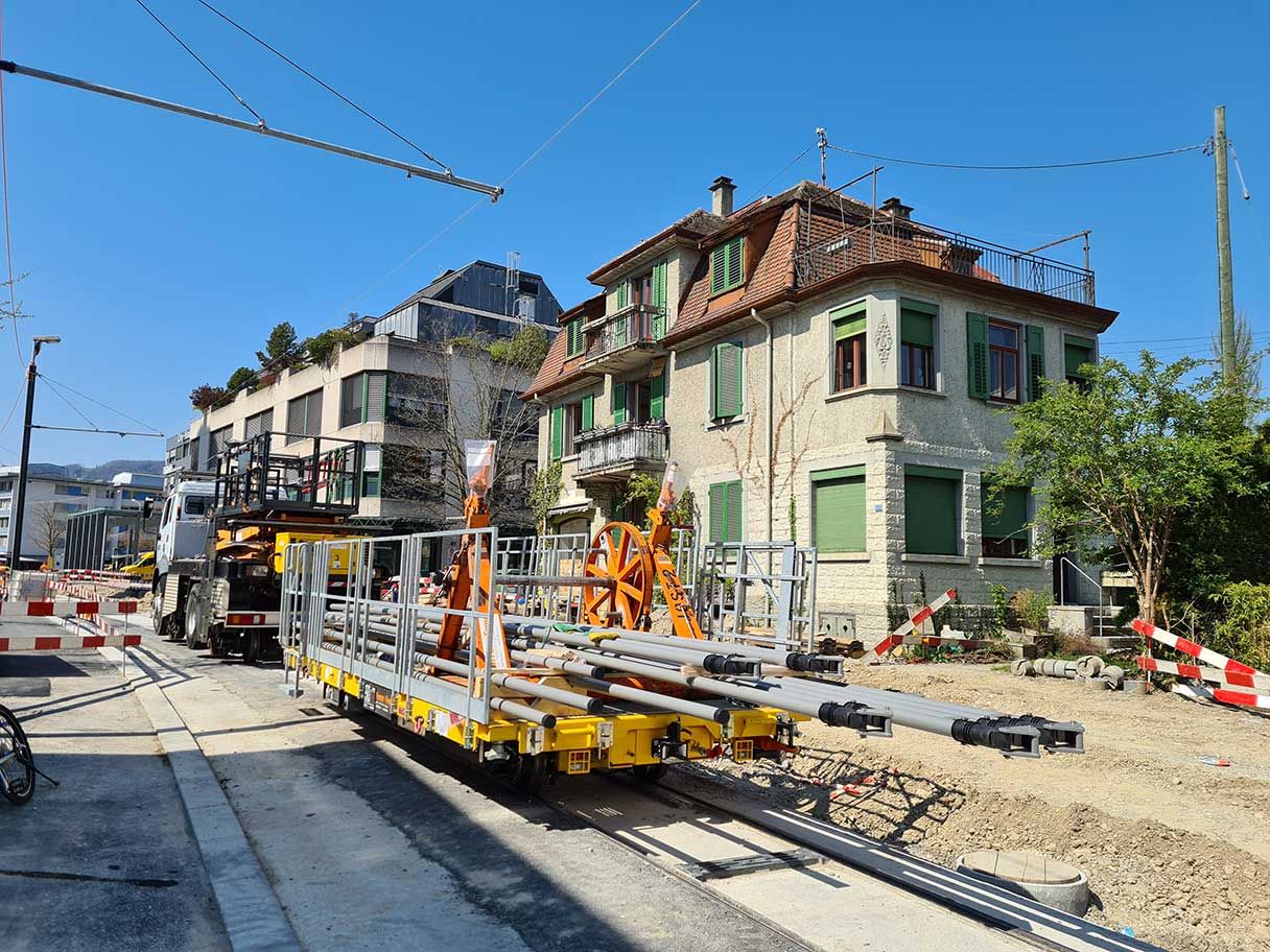 Eine Baustelle an einem sonnigen Tag mit mehreren Gebäuden im Hintergrund. Arbeiter scheinen Masten auf einem Flachbett-Schienenfahrzeug zu verlegen oder zu transportieren. Entlang der Straße befinden sich Bauzäune, Geräte und Maschinen.