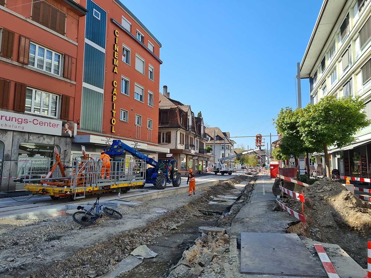 Eine Straße im Bau, in der Arbeiter in orangefarbenen Uniformen graben und Arbeiten an schweren Maschinen durchführen. Der Bereich ist mit Absperrungen abgesperrt, und im Hintergrund links ist ein Gebäude mit der Aufschrift „Cinema Capitol“ zu sehen.
