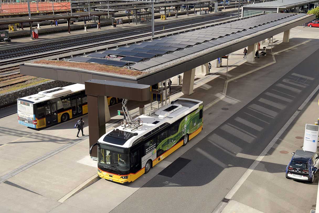 Eine Draufsicht auf einen Busbahnhof mit Elektrobussen, die unter einer Solarpanel-Struktur geparkt sind. Einige Passagiere laufen herum und im Hintergrund sind Bahngleise zu sehen. Ein Bus ist grün und gelb, ein anderer weiß und gelb.