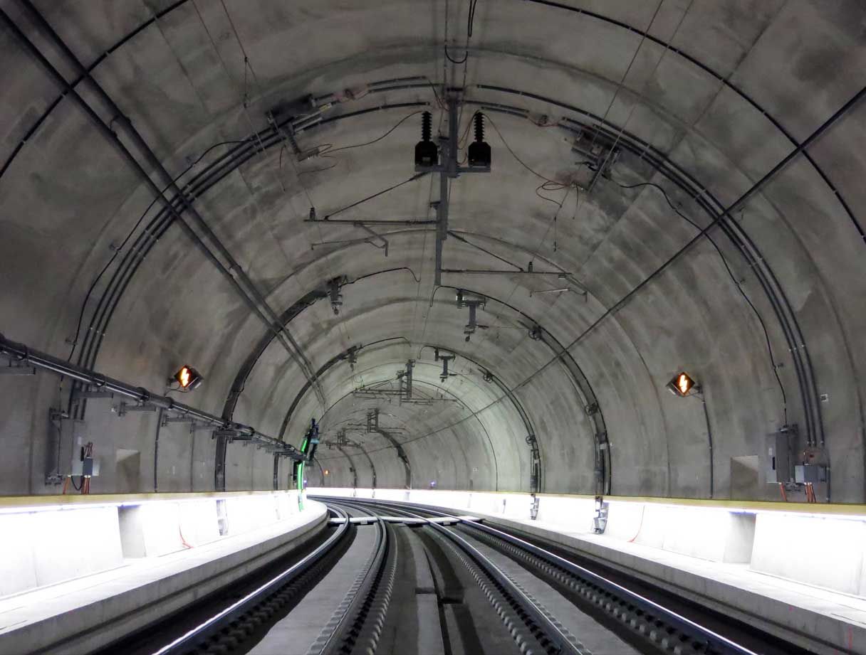 Ein gut beleuchteter, zweigleisiger Eisenbahntunnel aus Beton, der leicht nach links abknickt. Tunneldecke und -wände sind mit verschiedenen Kabeln und Geräten ausgekleidet. Die Gleise liegen auf Betonschwellen und der Boden des Tunnels ist sauber und frei von Schutt.
