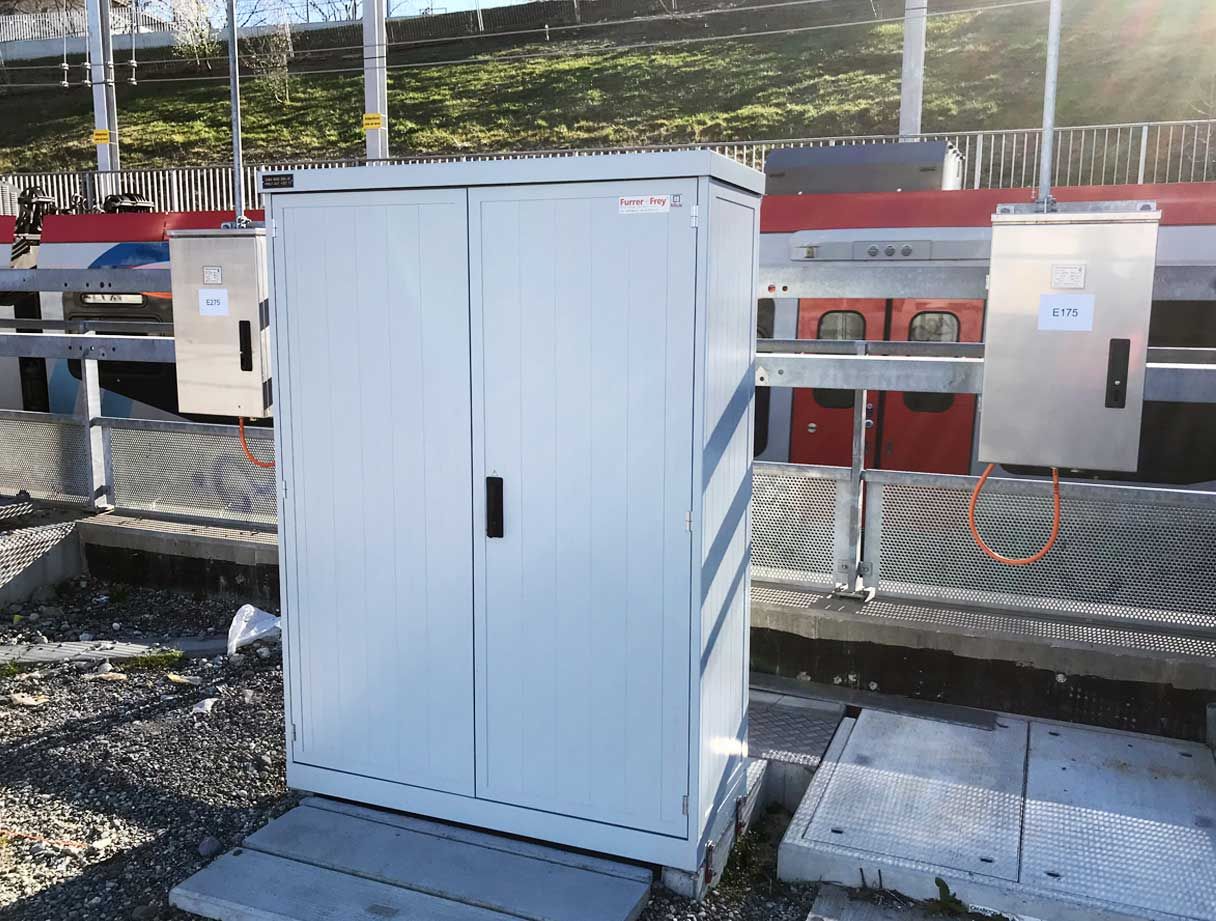 Auf einer Betonplattform steht ein grauer Metallschrank neben zwei kleineren Schaltkästen mit der Beschriftung E15 und E75. Dahinter sind ein roter Zug und Hochbahngleise zu sehen. Der Bereich ist von Geländern und verschiedenen Versorgungseinrichtungen umgeben.