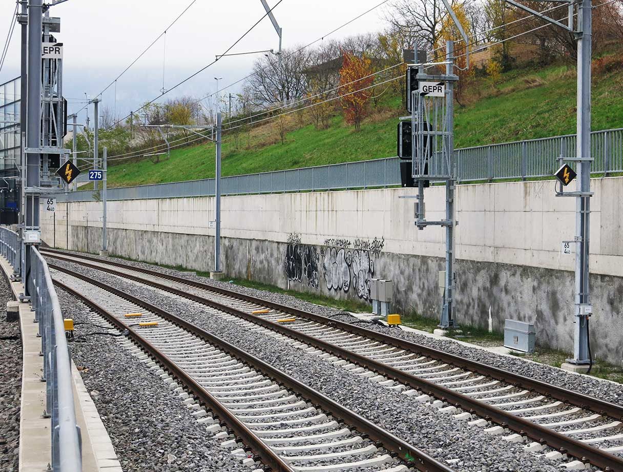 Ein Abschnitt von Eisenbahnschienen mit Oberleitungen neben einer mit Graffiti bedeckten Betonwand. Entlang der Gleise befinden sich Signallichter und -geräte, und hinter der Wand ist ein grasbewachsener Hügel zu sehen. Der Himmel ist bedeckt.