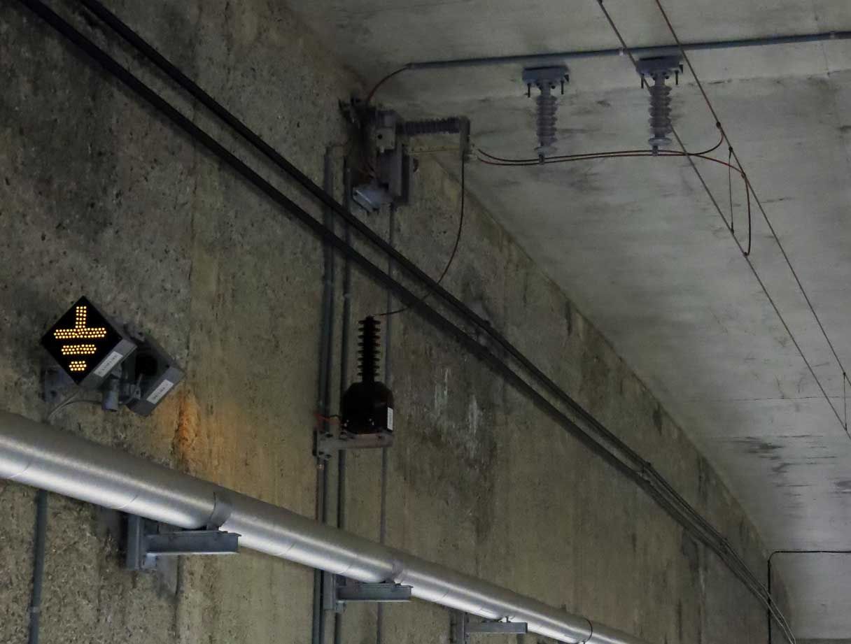 Eine Tunneldecke aus Beton, die mit verschiedenen elektrischen und mechanischen Komponenten ausgestattet ist, darunter Isolatoren, Kabel und Anschlusskästen. Auf der linken Seite der Wand ist ein kleines beleuchtetes Schild mit einem nach unten zeigenden Pfeil angebracht.
