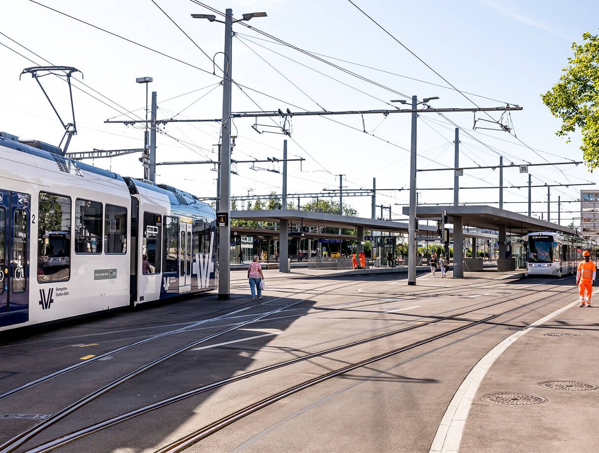 Eine moderne Straßenbahnhaltestelle mit mehreren Straßenbahnen, die auf den Gleisen halten. Menschen laufen auf dem Bahnsteig herum und ein paar Arbeiter in orangefarbenen Sicherheitsuniformen sind zu sehen. Die Szene ist hell mit einem klaren Himmel und mehreren Oberleitungen, die sich darüber kreuzen.