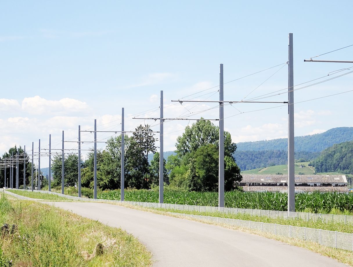 Eine Landstraße, die auf beiden Seiten von Gras und Vegetation gesäumt ist und zu Reihen hoher Strommasten führt, über die Leitungen verlaufen. Im Hintergrund sind üppige grüne Bäume und in der Ferne sanfte Hügel unter einem teilweise bewölkten Himmel zu sehen.