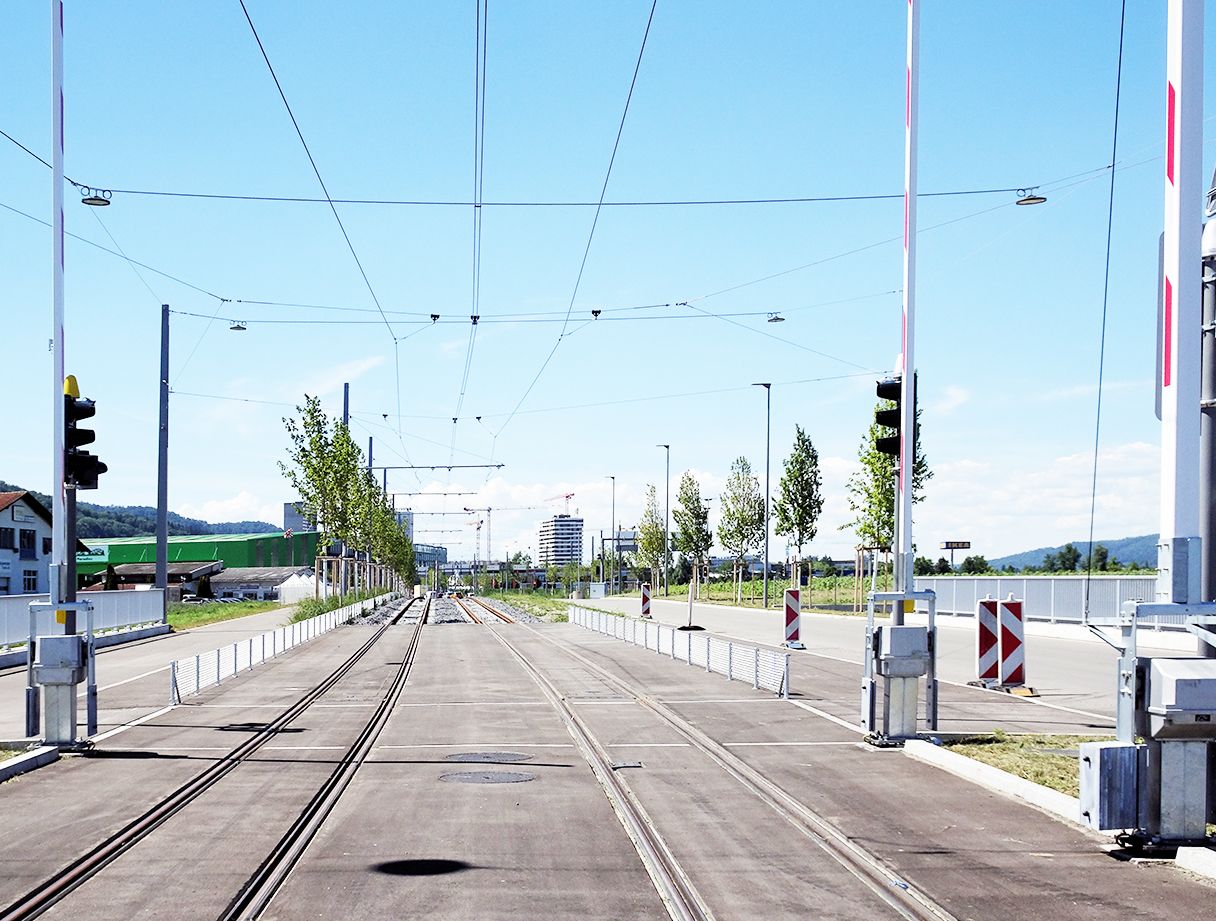 An einem hellen, klaren Tag erstreckt sich in der Ferne eine moderne Straßenbahn mit Gleisen, Ampeln und Oberleitungen. Bäume säumen beide Seiten der Gleise, und im Hintergrund sind neben einem klaren blauen Himmel Gebäude zu sehen. An den Seiten befinden sich Straßenbarrieren.