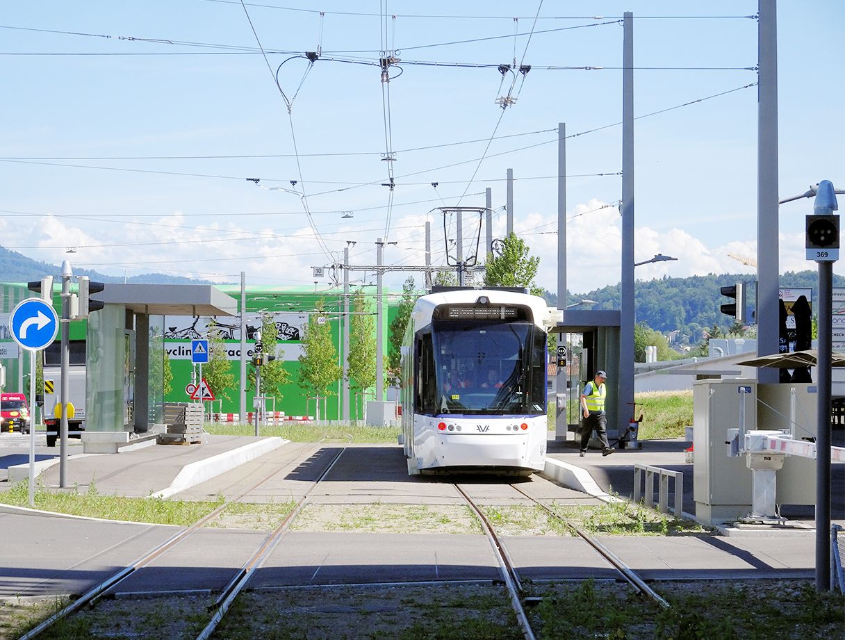 Eine weiße Straßenbahn nähert sich einer modernen Straßenbahnhaltestelle mit Oberleitungen. Zwei Personen stehen auf dem Bahnsteig: eine in Uniform neben der Straßenbahn und eine andere wartend. Die Umgebung ist städtisch mit blauem Himmel und vereinzelten Wolken im Hintergrund.