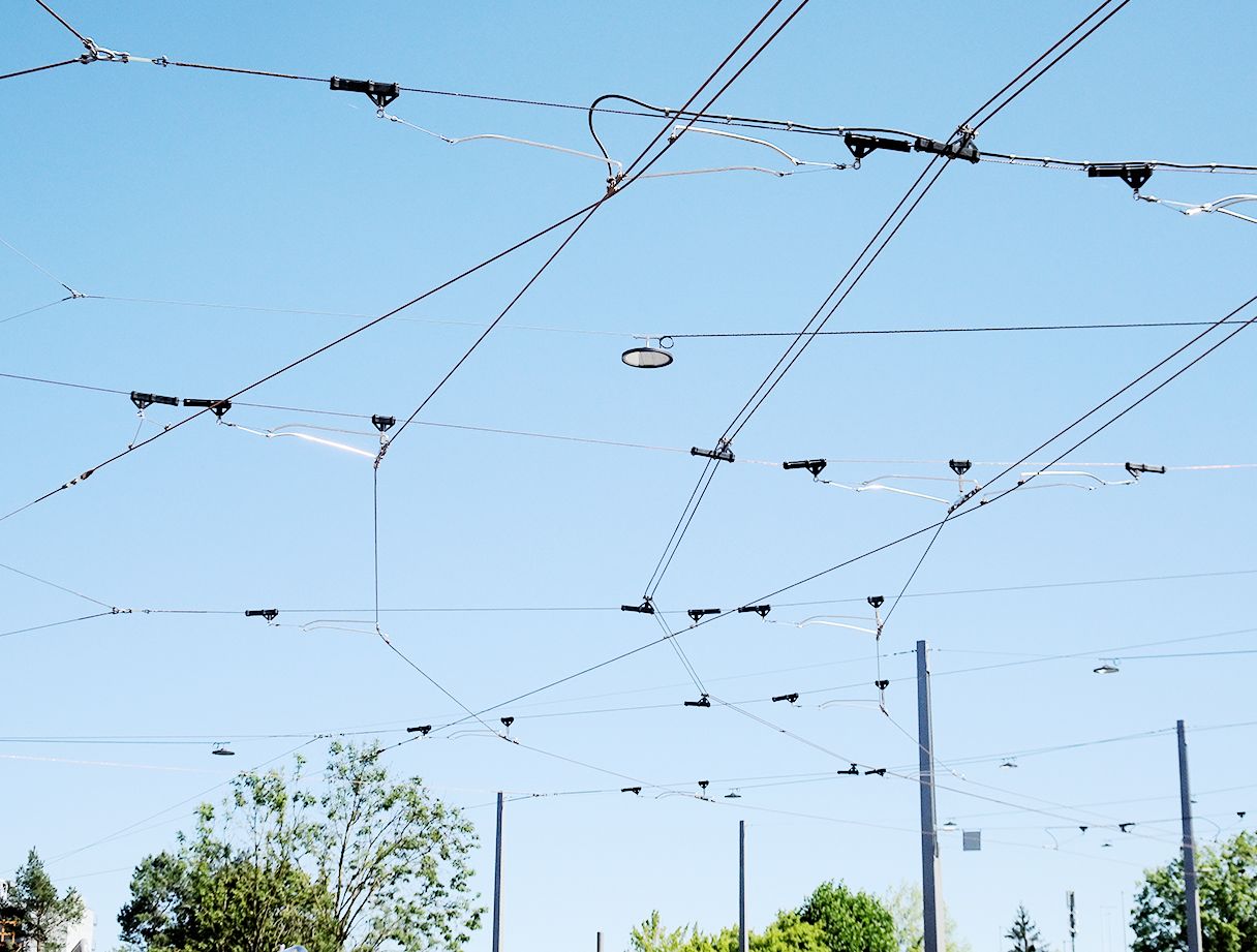 Ein klarer blauer Himmel mit einem Netz aus kreuz und quer verlaufenden Straßenbahn- oder Trolleybusleitungen. Hohe Masten stützen die Leitungen und im unteren Teil des Bildes sind einige Bäume und Blätter zu sehen.