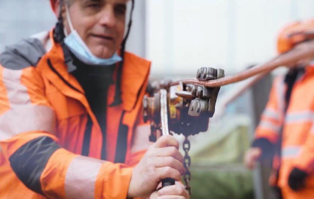 Ein Arbeiter in einer orangefarbenen Sicherheitsweste bedient im Freien schwere Maschinen. Er hält einen an der Maschine befestigten Hebel, während ein anderer Arbeiter in Orange verschwommen im Hintergrund zu sehen ist. Der Arbeiter trägt einen Schutzhelm und hat eine Gesichtsmaske bis unter sein Kinn heruntergezogen.
