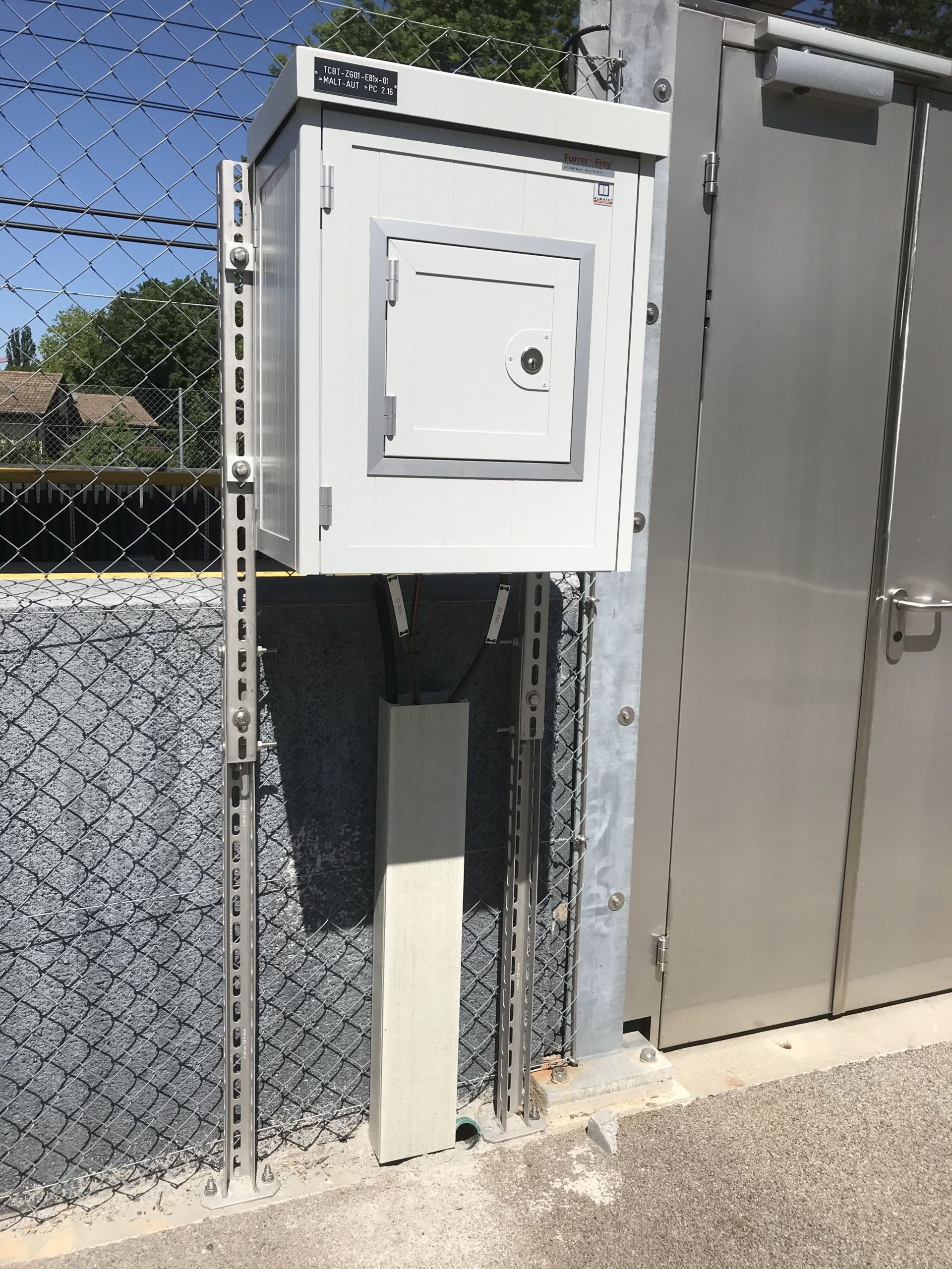 Ein kleiner, weißer Metall-Schaltkasten, der an einem Zaun montiert ist und an dessen Tür ein Schloss angebracht ist. Er steht zwischen Metallpfosten und neben einer Edelstahltür. Der Zaun besteht aus Maschendraht mit grauer Basis und in der Umgebung befinden sich Bäume und ein klarer, sonniger Himmel.