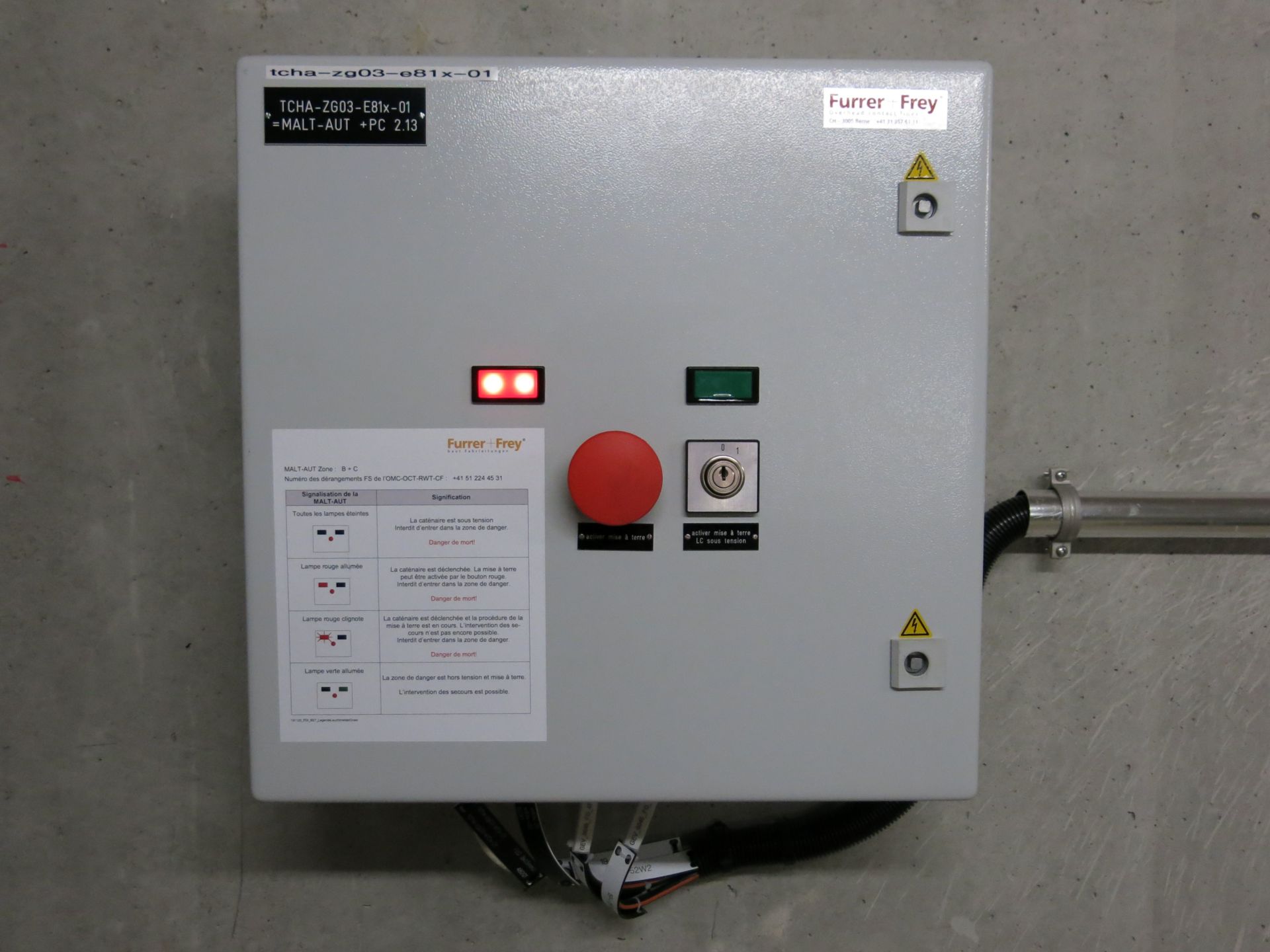 Ein graues Bedienfeld mit verschiedenen Warnhinweisen, einem großen roten Not-Aus-Schalter, einem grünen Startknopf, einem Schlüsselschalter und einem informativen Hinweisschild auf der Vorderseite. Das Bedienfeld ist an einer Betonwand montiert und auf der rechten Seite mit einem Kabel ausgestattet.