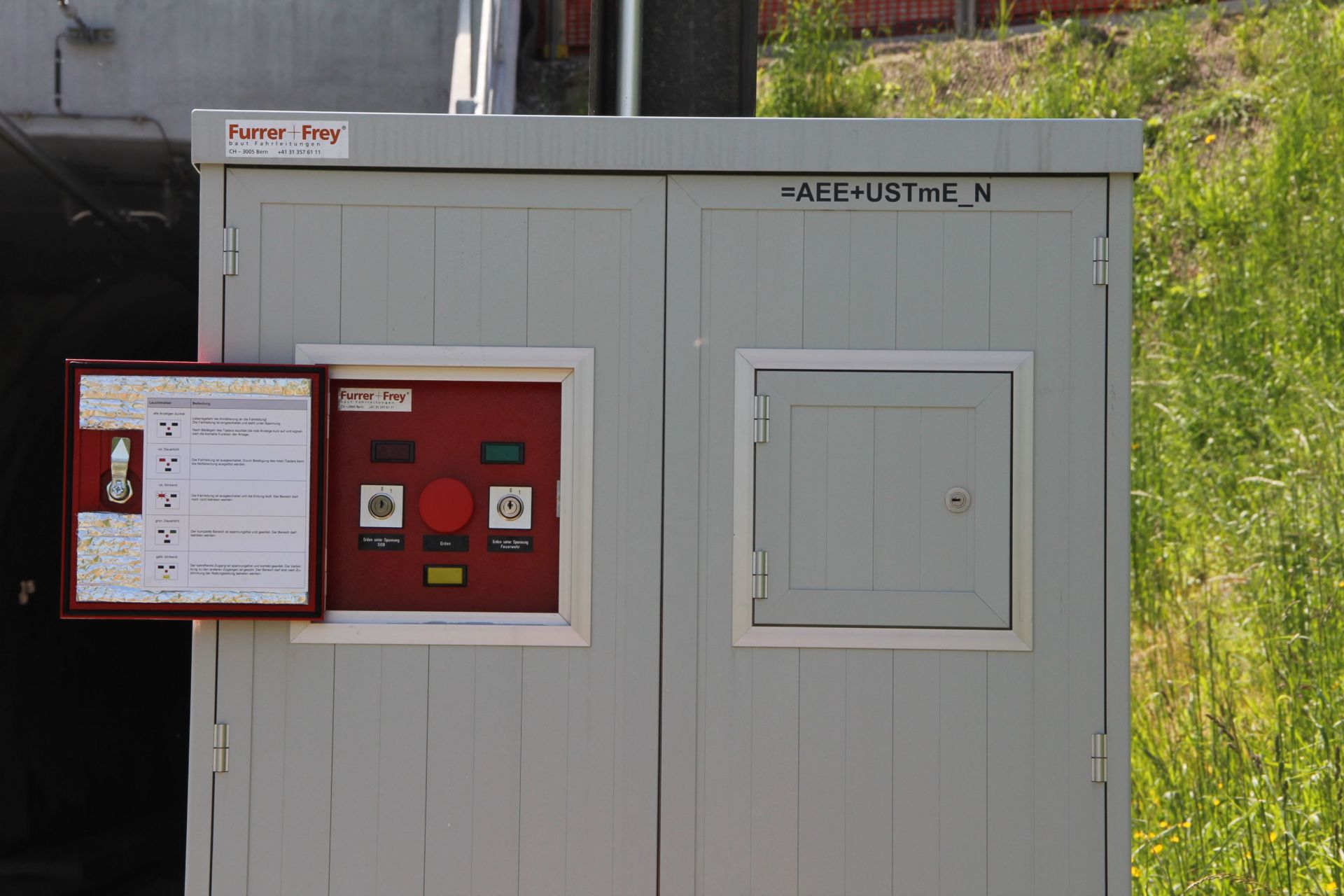 Ein graues Bedienfeld im Außenbereich mit grünem Hintergrund. Es verfügt über mehrere Knöpfe und Anzeigen. Außerdem gibt es ein kleines Schild mit Anweisungen und einen roten Knopf. Oben auf dem Bedienfeld sind „Furrer + Frey“ und einige Buchstaben/Zahlen zu sehen.