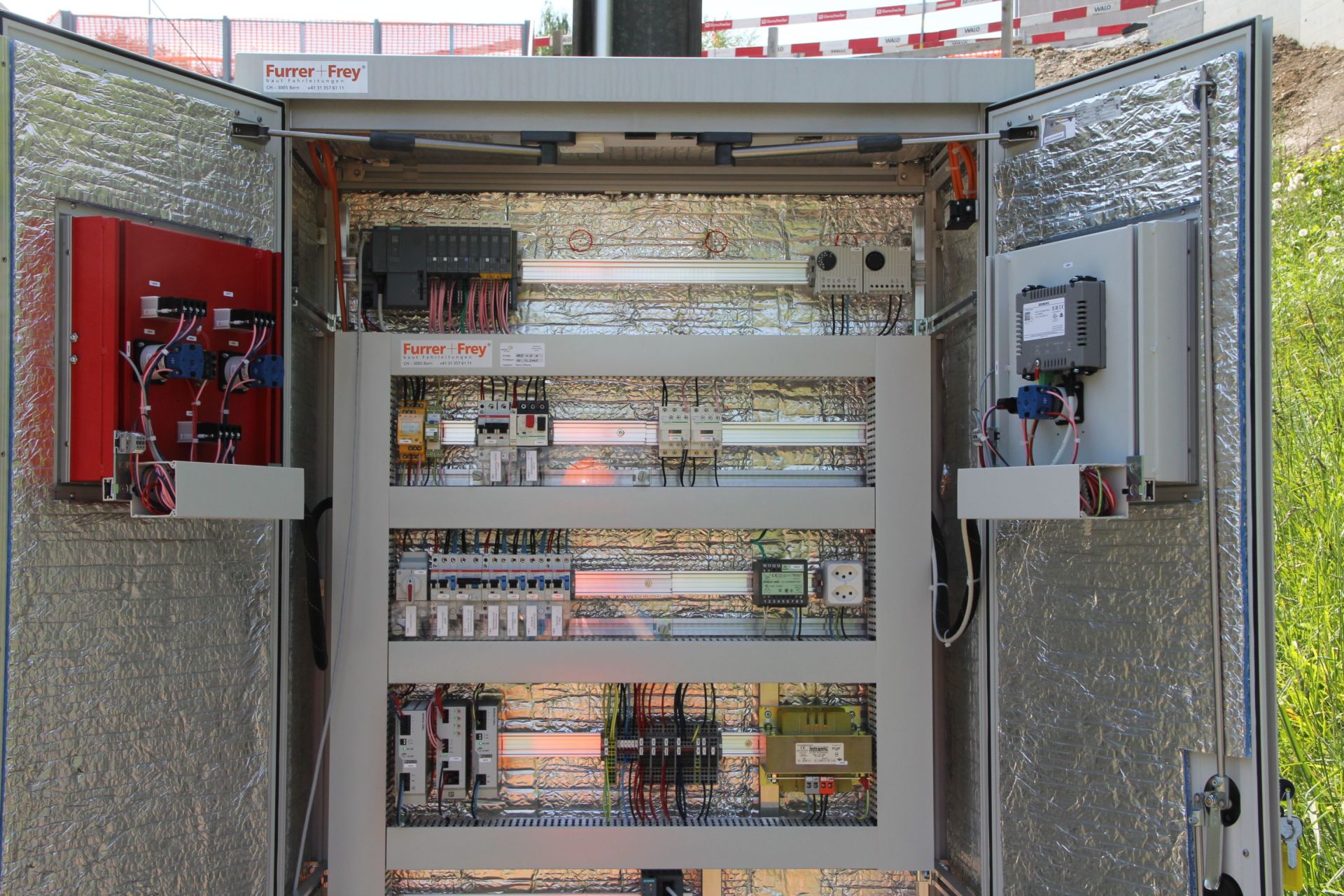 Ein offener Schaltschrank, in dem verschiedene installierte elektrische Komponenten, Kabel und Geräte auf Paneelen im Inneren zu sehen sind. Die Schranktüren sind geöffnet und geben den Blick auf Kabel und Geräte frei, mit Beschriftungen und einer metallisch reflektierenden Innenfläche.