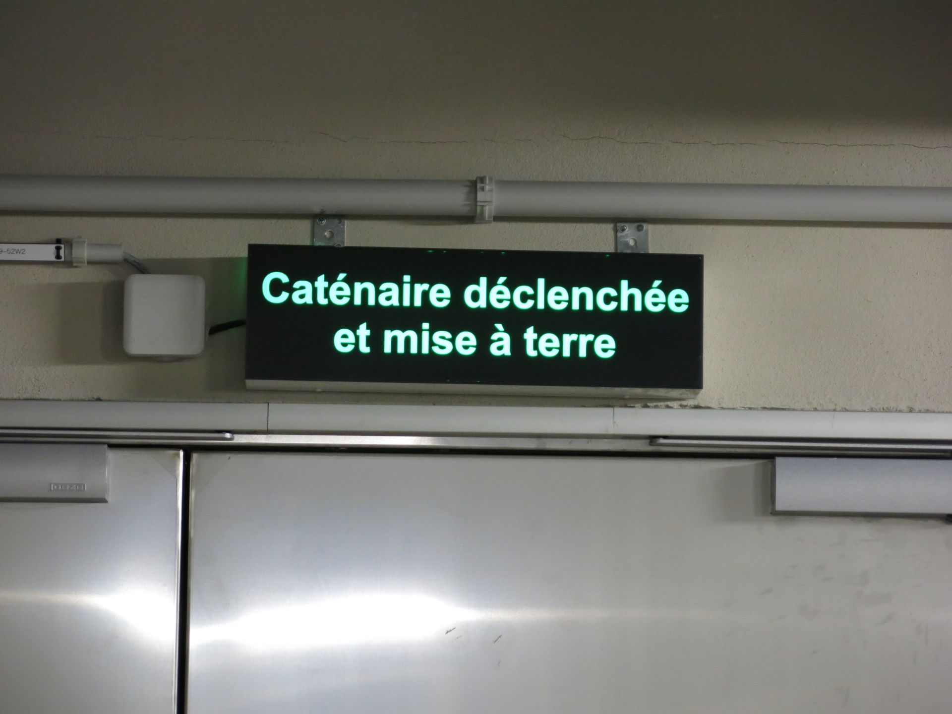 An einer Wand über einer Metalltür ist ein Schild mit dem französischen Text „Caténaire déclenchée et mise à terre“ angebracht. Der Text auf dem Schild ist weiß und leuchtet auf schwarzem Hintergrund.