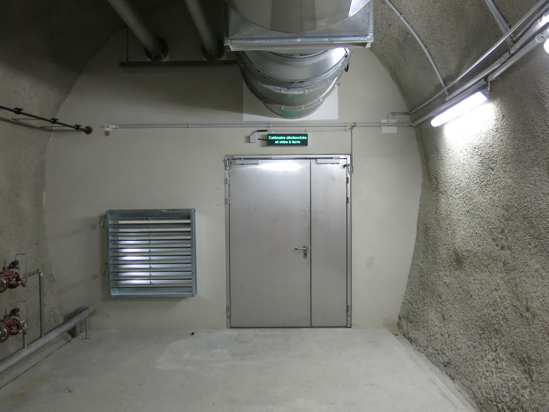 Ein schlichter Industriekorridor mit Betonwänden führt zu einer Doppeltür aus Metall unter einem grünen Ausgangsschild mit der Aufschrift „Kontrollraum“. An der Decke verlaufen Rohre, und in die linke Wand ist eine große Lüftungsöffnung eingelassen. Leuchtstofflampen sorgen für die Beleuchtung.