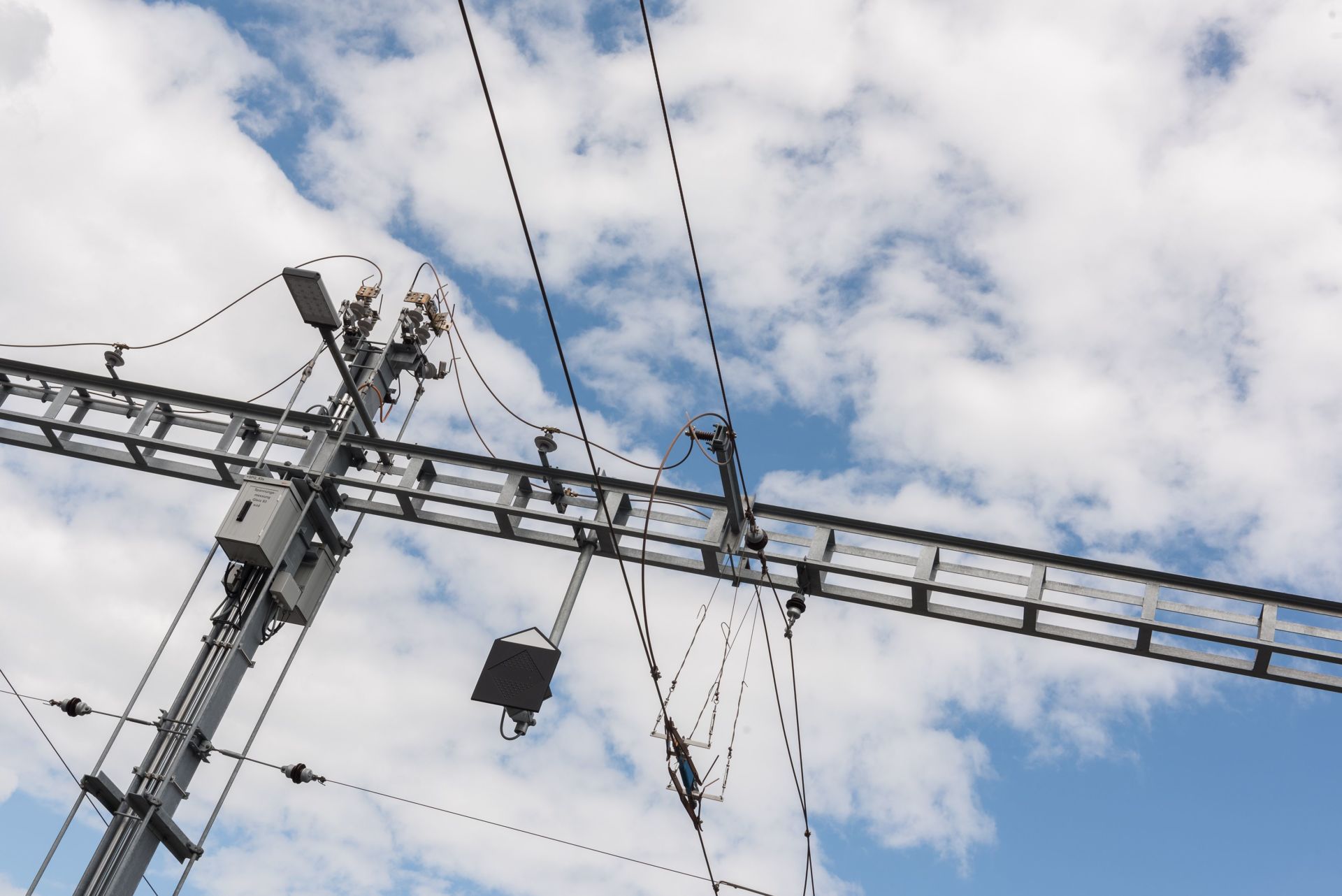 Oberirdische elektrische Leitungen und Infrastruktur vor einem teilweise bewölkten Himmel. Das Bild zeigt Metallträgerstrukturen, Isolatoren und verschiedene elektrische Komponenten. Diese Anordnung wird typischerweise für die Stromversorgung elektrischer Züge oder Straßenbahnen verwendet.