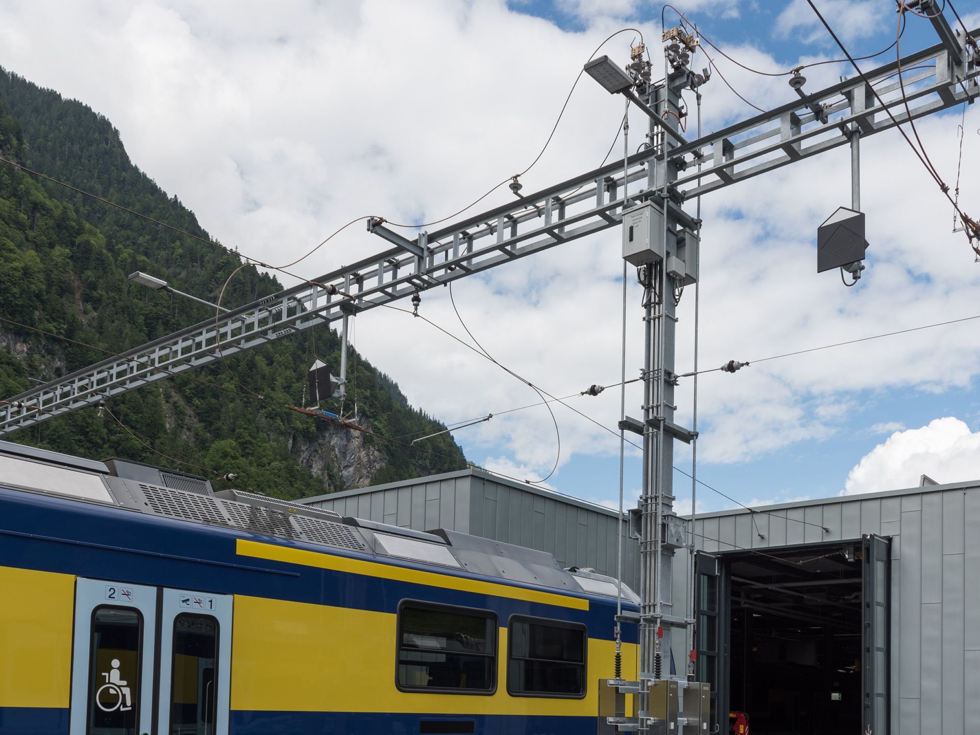 Ein blau-gelber Zug steht neben einem Bahnhofsgebäude mit elektrischen Oberleitungen und -strukturen. Im Hintergrund sind ein mit grünen Bäumen bedeckter Berg und ein wolkiger Himmel zu sehen. Das Gebäude hat einen offenen, garagenartigen Eingang.