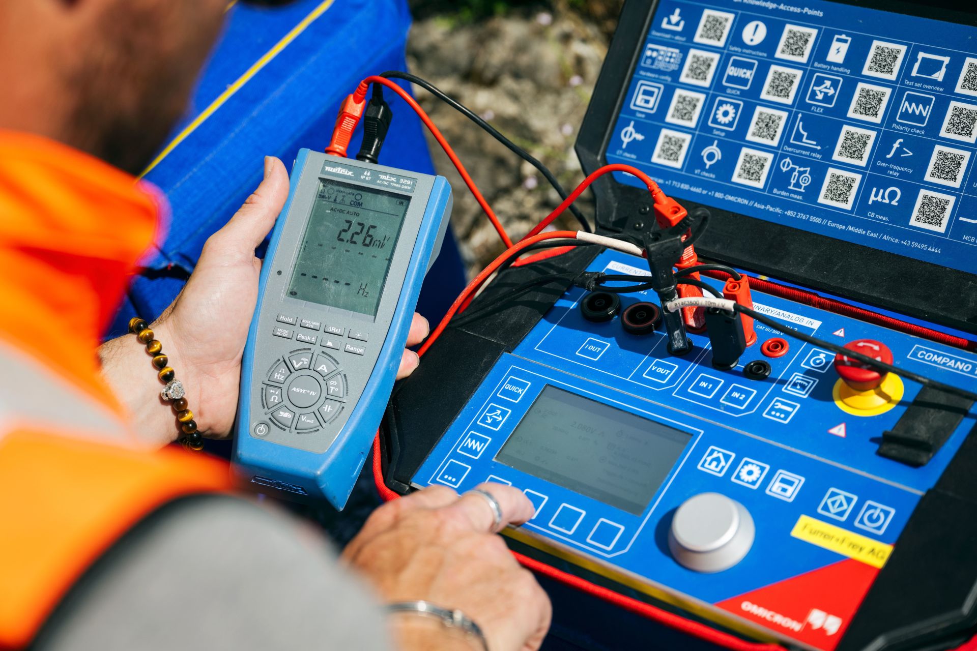 Eine Person mit einer orangefarbenen Sicherheitsweste bedient ein elektronisches Prüfgerät. Sie hält ein Handgerät in der Hand, das über Kabel mit einem anderen Gerät verbunden ist, das über einen Bildschirm, Knöpfe, Skalen und Referenzsymbole verfügt.