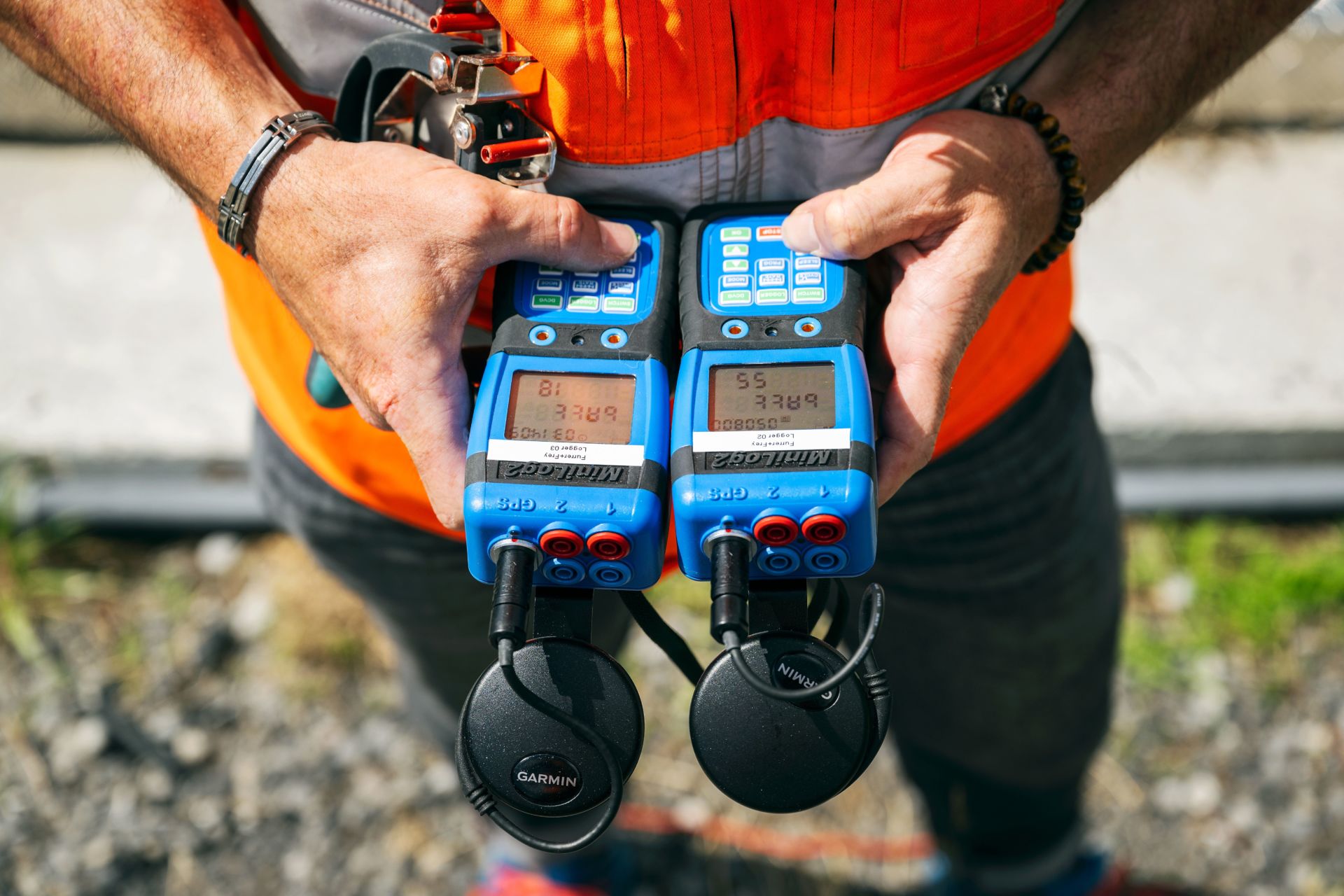 Eine Person mit einer orangefarbenen Sicherheitsweste hält zwei blaue elektronische Geräte mit Zahlenanzeigen in der Hand. An den Geräten sind Kopfhörer mit der Aufschrift „Garmin“ angebracht. Die Person steht im Freien auf Kies, im Hintergrund ist grünes Gras zu sehen.