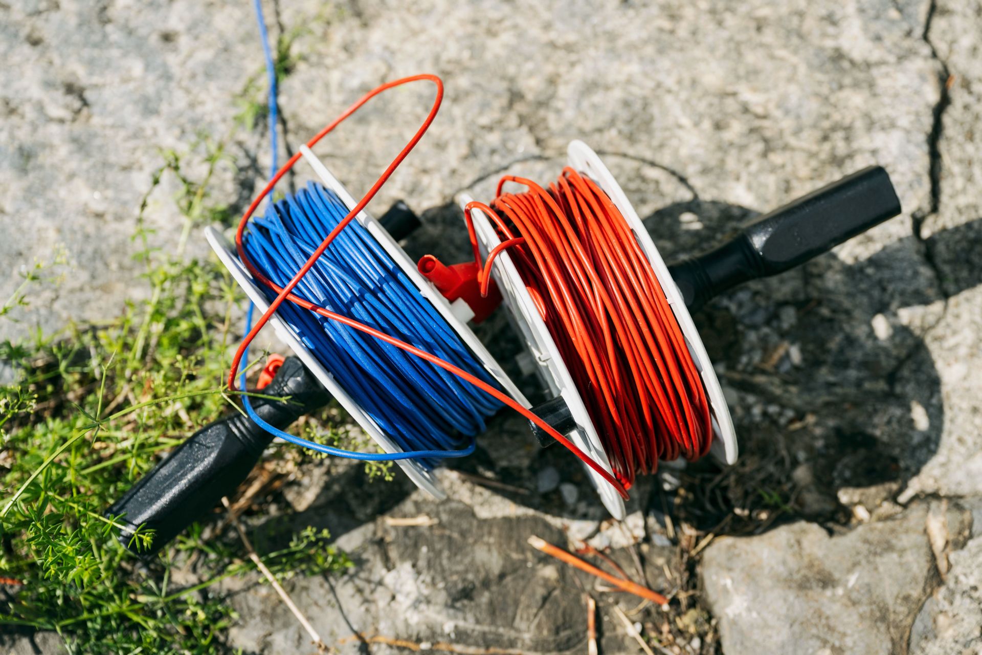 Auf dem Boden liegen zwei Drahtspulen, eine mit blauem Draht und die andere mit rotem Draht. Die Spulen wickeln sich ab, und einige Drähte sind verheddert. Der Boden ist rau und mit Schutt und einem kleinen Fleck grünem Gras übersät.