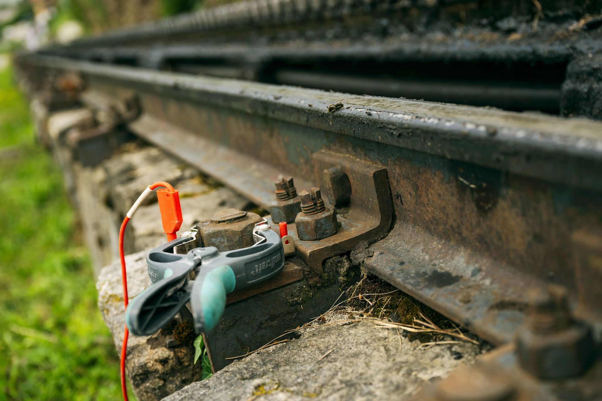 Nahaufnahme eines rostigen Eisenbahngleises mit einem daran mit einer Klammer befestigten elektrischen Gerät. Das Gerät hat rote und weiße Drähte und das Gleis ist mit sichtbaren Schrauben und Muttern auf einer Betonplatte montiert. Um die Platte herum wächst Gras.