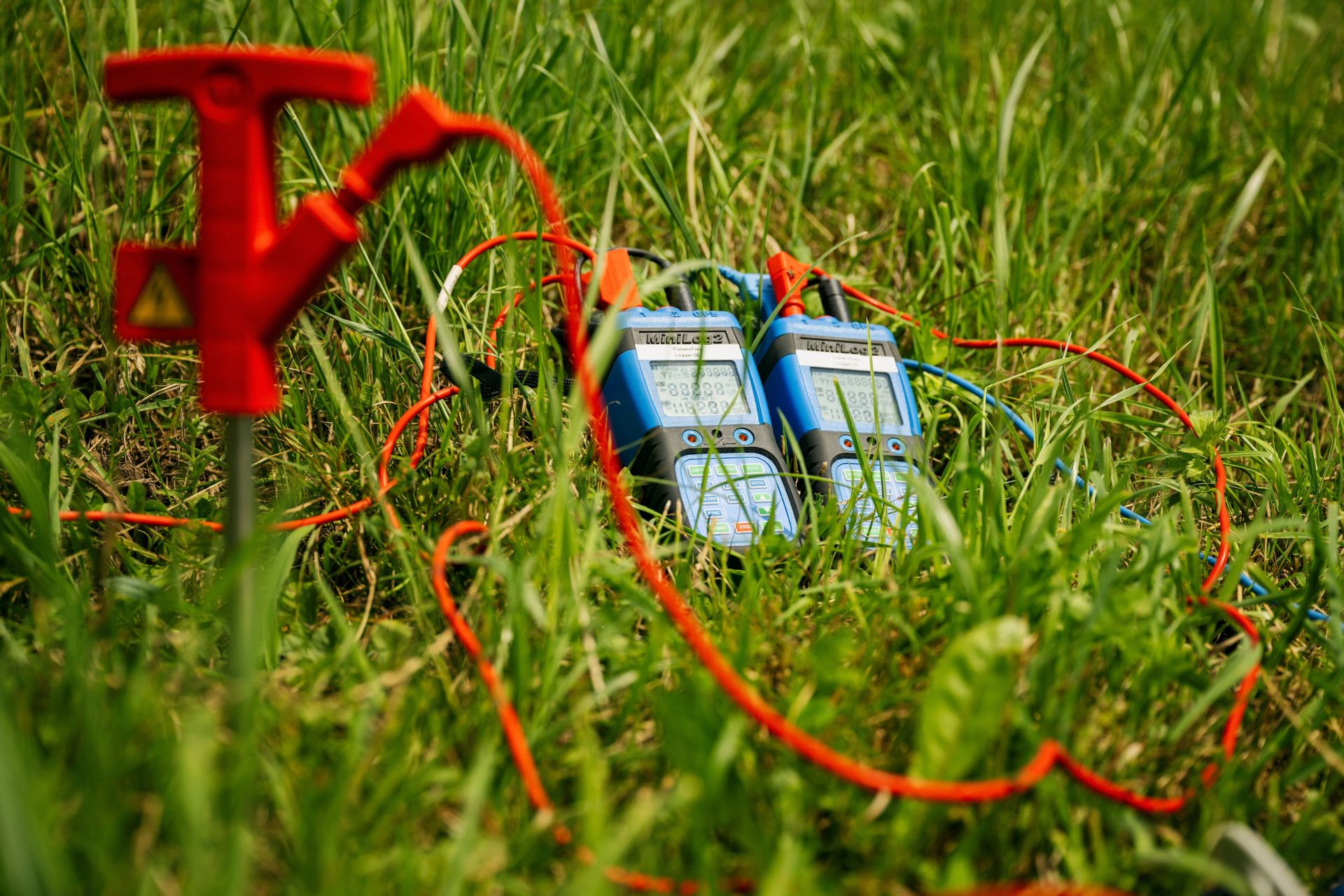 Zwei elektronische Geräte mit Bildschirmen und mehreren angeschlossenen roten Kabeln liegen auf dem Rasen. Auf der linken Seite ist ein roter Erdspieß zu sehen. Der Aufbau scheint für eine Feldmessung oder Testaktivität zu sein.