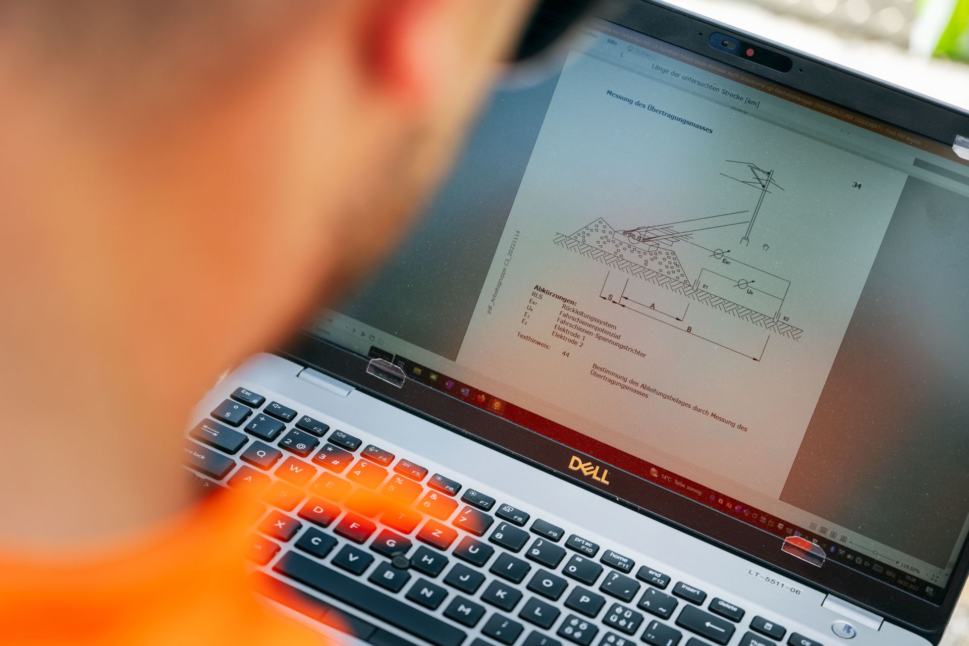 Eine Person in einem orangefarbenen Hemd betrachtet ein technisches Dokument auf einem Dell-Laptop. Das auf dem Bildschirm sichtbare Dokument enthält ein detailliertes Diagramm und Messungen, was auf ein technisches oder architektonisches Thema schließen lässt. Die Person ist von hinten zu sehen und konzentriert sich auf den Bildschirm.