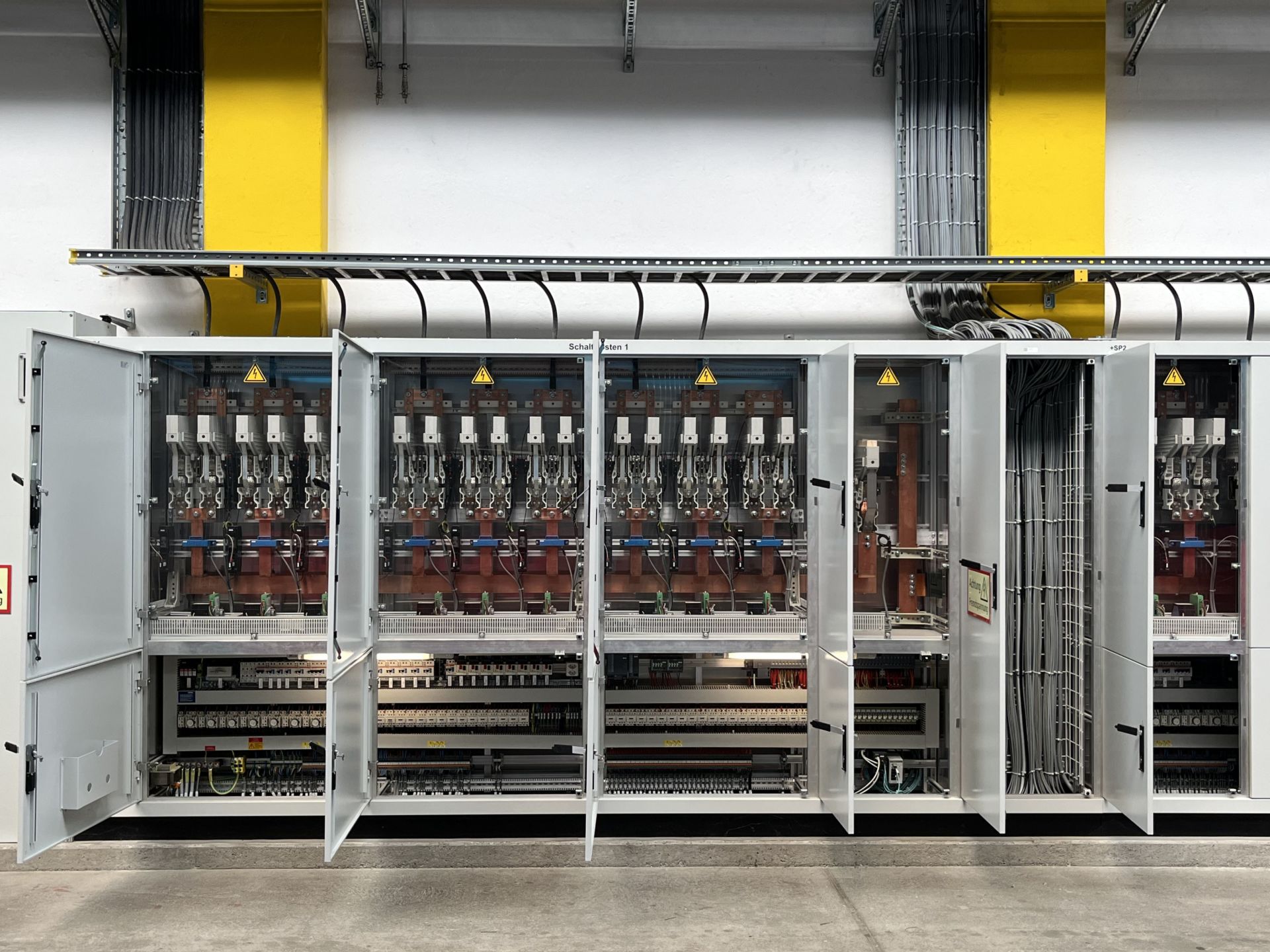 Ein offener Schaltschrank mit mehreren breiten Türen, hinter denen eine komplexe Anordnung von elektrischen Komponenten, Schaltern und Kabeln zu sehen ist. Der Schaltschrank ist systematisch aufgebaut und befindet sich in einem Gebäude mit einer gelb-weißen Wand und Oberleitung.