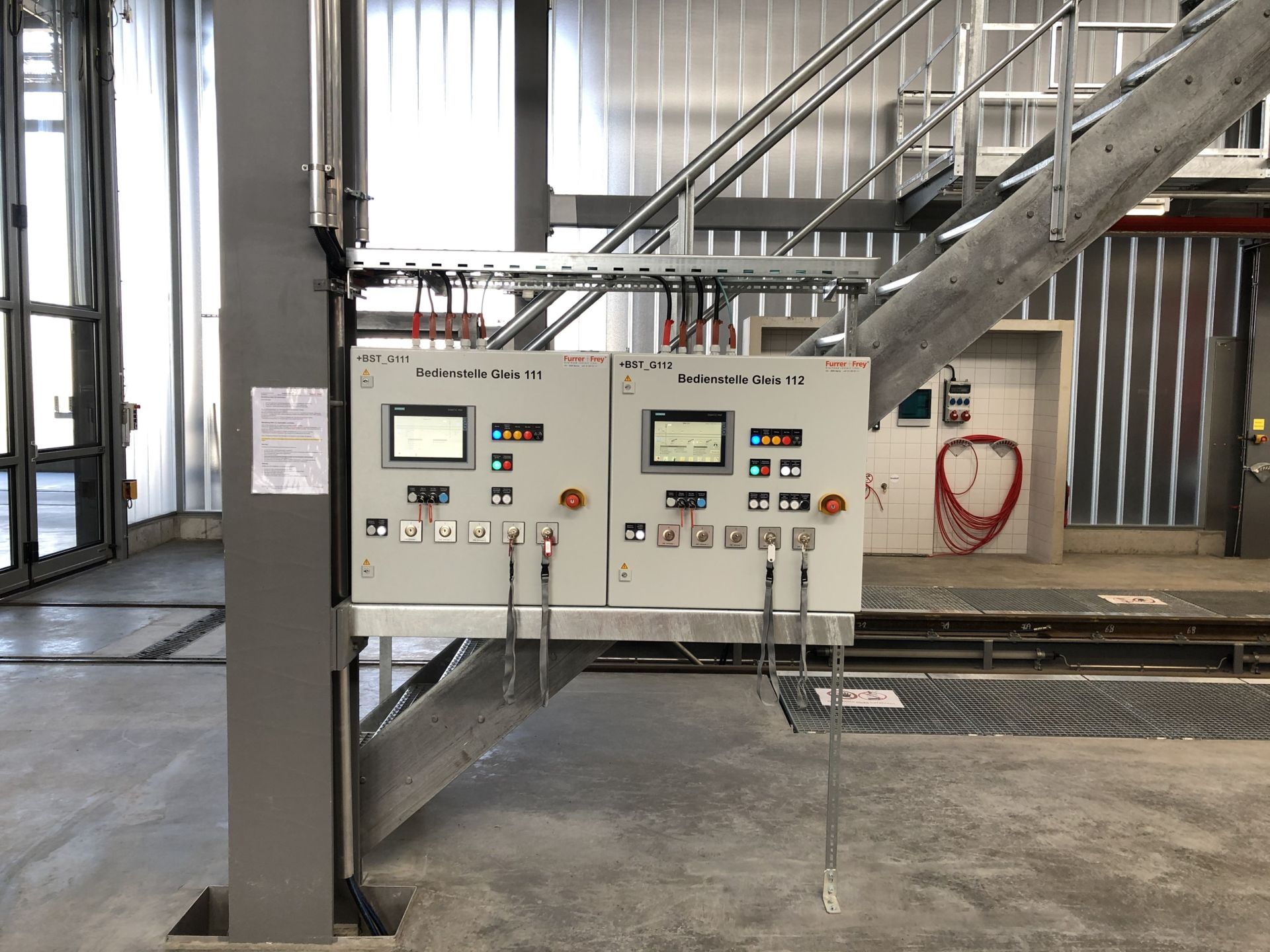 Ein Bedienfeld mit mehreren Tasten und digitalen Anzeigen ist auf einer Metallkonstruktion in einer Industrieanlage montiert. Das Bedienfeld befindet sich unter einer Metalltreppe mit Geländer. Im Hintergrund hängen rote Schläuche an einer Wand. Der Boden ist aus Beton.