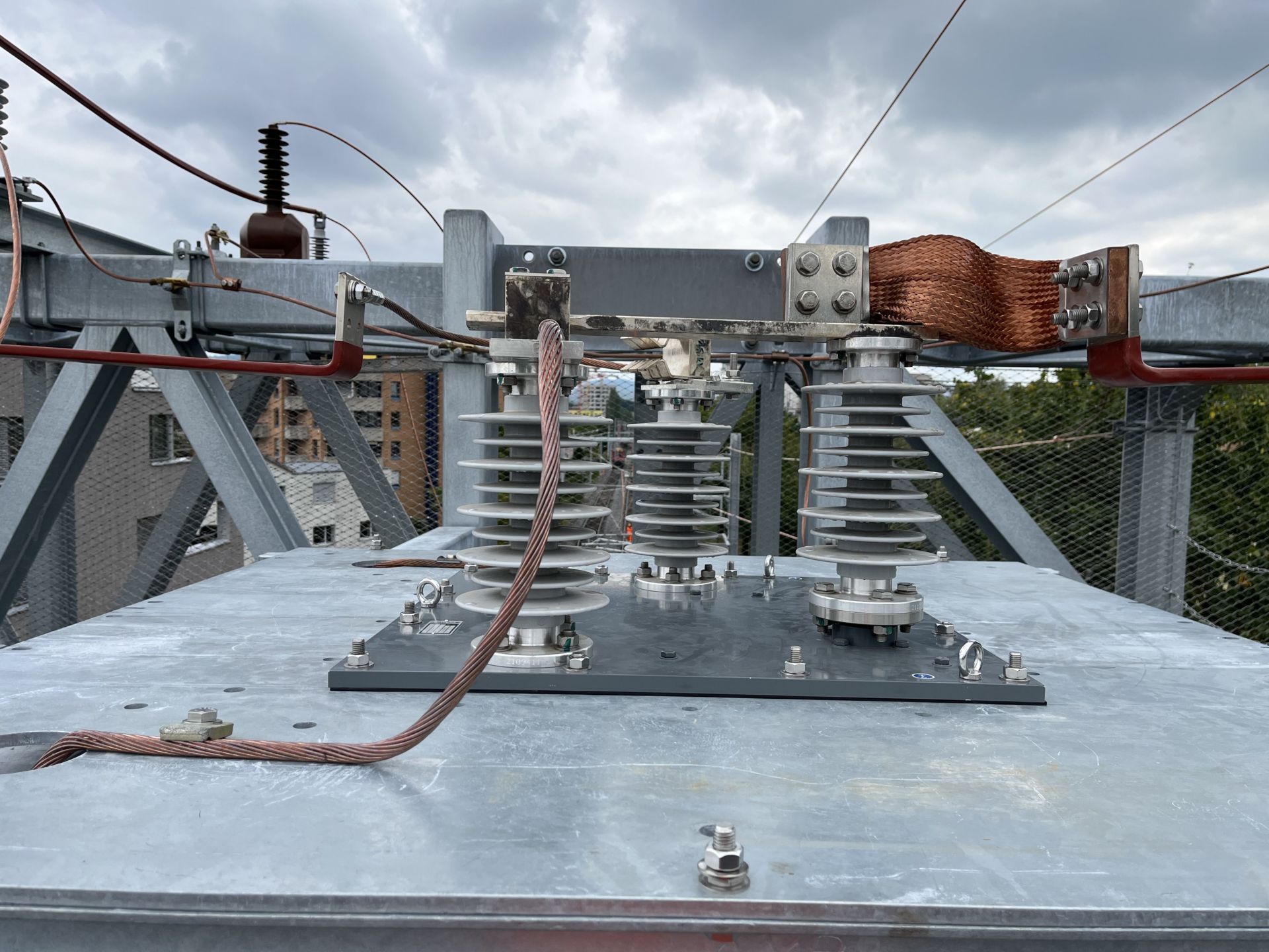 Nahaufnahme von elektrischen Isolatoren und Komponenten, die auf einer Metallstruktur montiert sind und an die Kabel angeschlossen sind. Die Anordnung scheint Teil eines Umspannwerks oder Stromverteilungssystems zu sein. Der Hintergrund ist bewölkt, Gebäude und Grünflächen sind sichtbar.