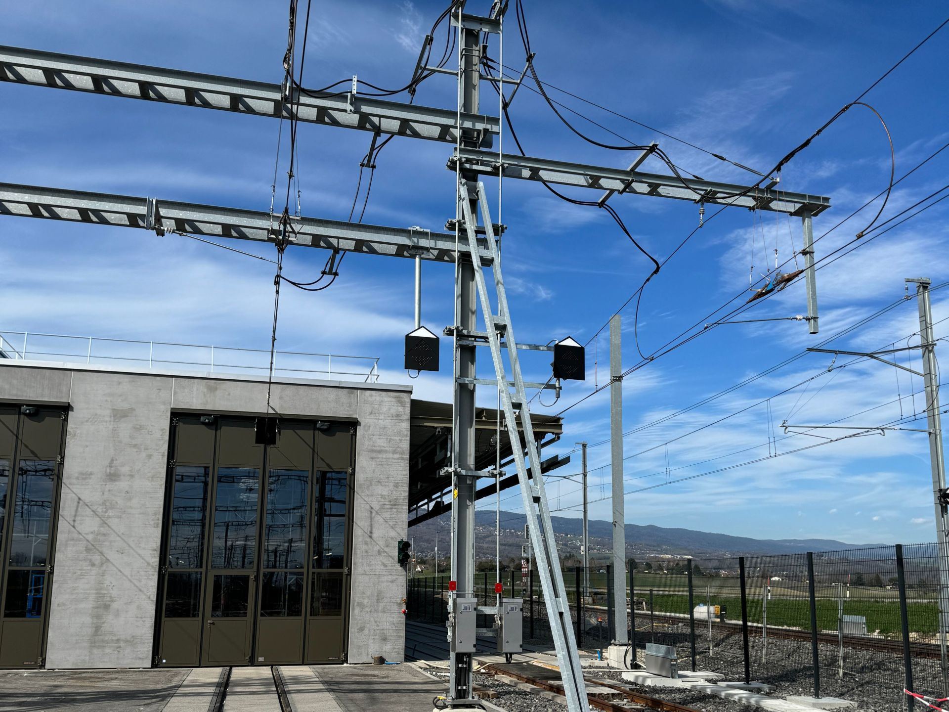 Eine Eisenbahninfrastrukturanlage mit Oberleitungen und Stützstrukturen vor einem Gebäude mit großen Türen. Im Hintergrund sind ein klarer blauer Himmel und Hügel in der Ferne zu sehen.