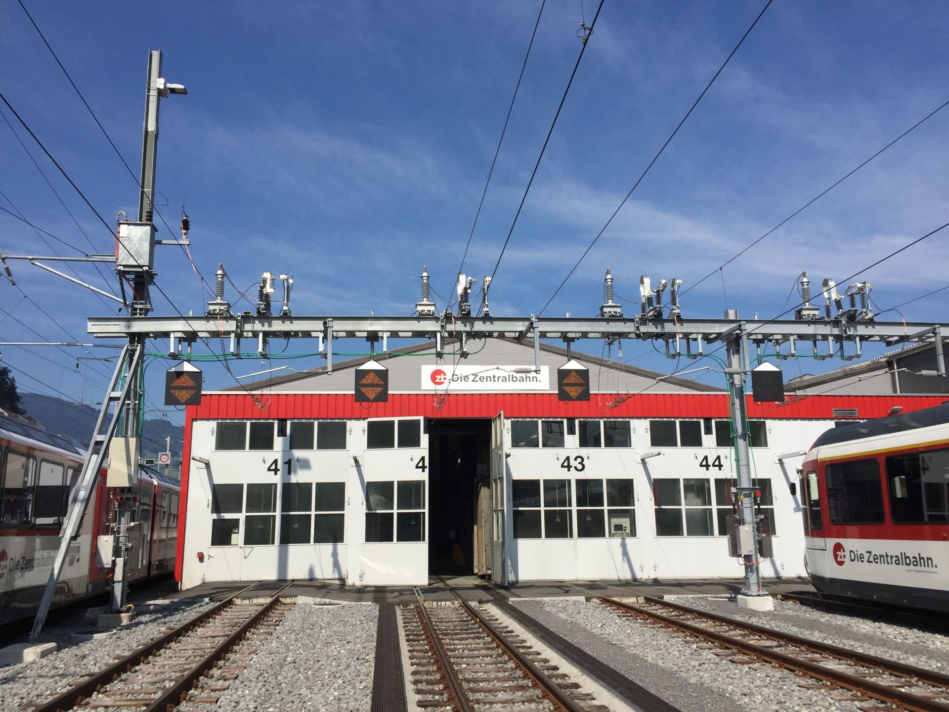 Ein Zugwartungsschuppen mit mehreren Gleisen, die zu ihm führen. Das Gebäude hat große weiße Türen mit der Aufschrift „43“ und „44“. Oberleitungen und Schienenausrüstung sind sichtbar, zusammen mit Zügen auf beiden Seiten, die mit dem Logo der „Zentralbahn“ gekennzeichnet sind.