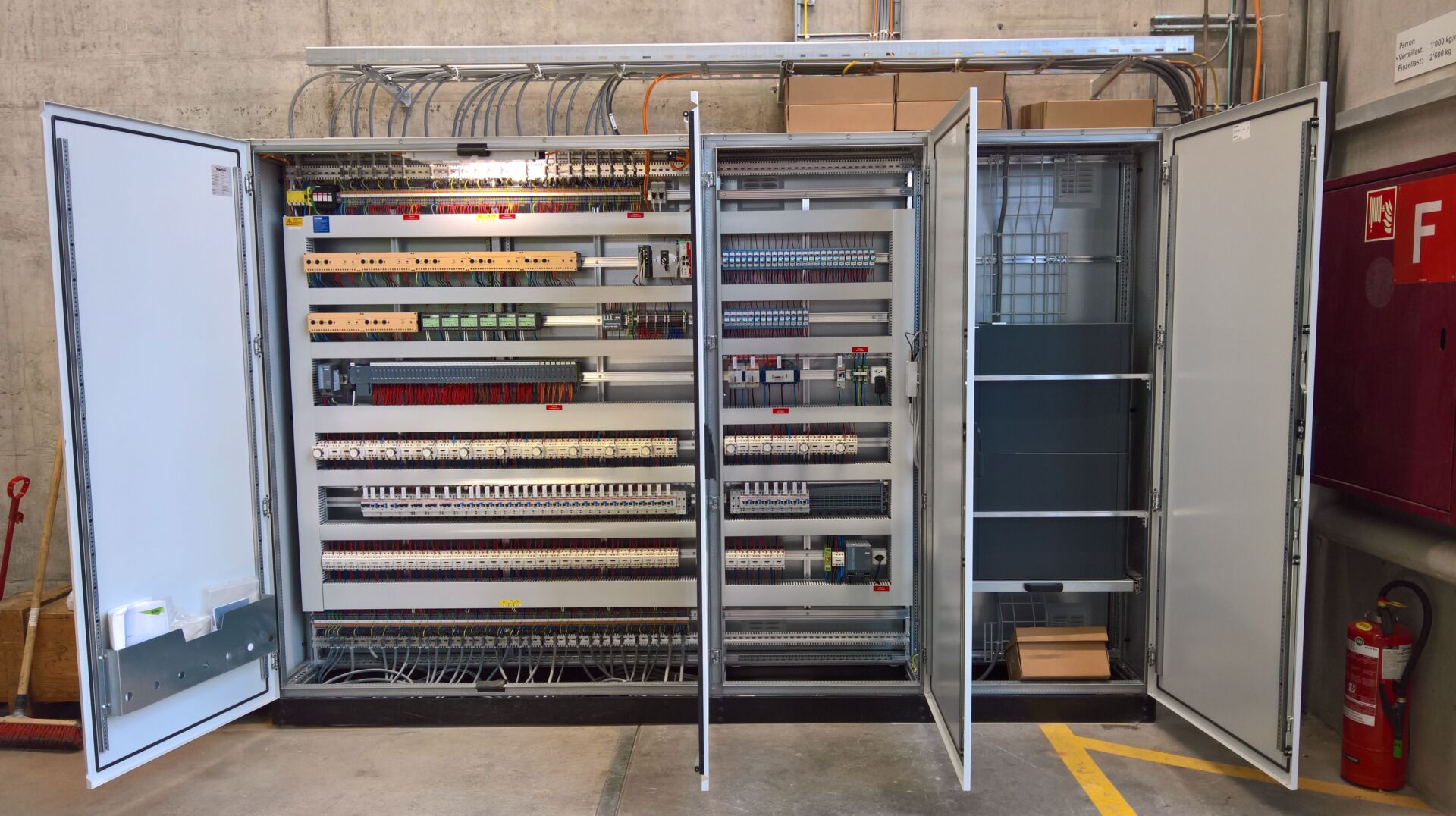 Ein offener, hoher Schaltschrank mit mehreren Abschnitten und Türen, die komplexe Anordnungen von Kabeln, Schaltern, Leistungsschaltern und elektronischen Komponenten offenbaren. Der Schrank steht an einer Betonwand in einer Industrieumgebung.