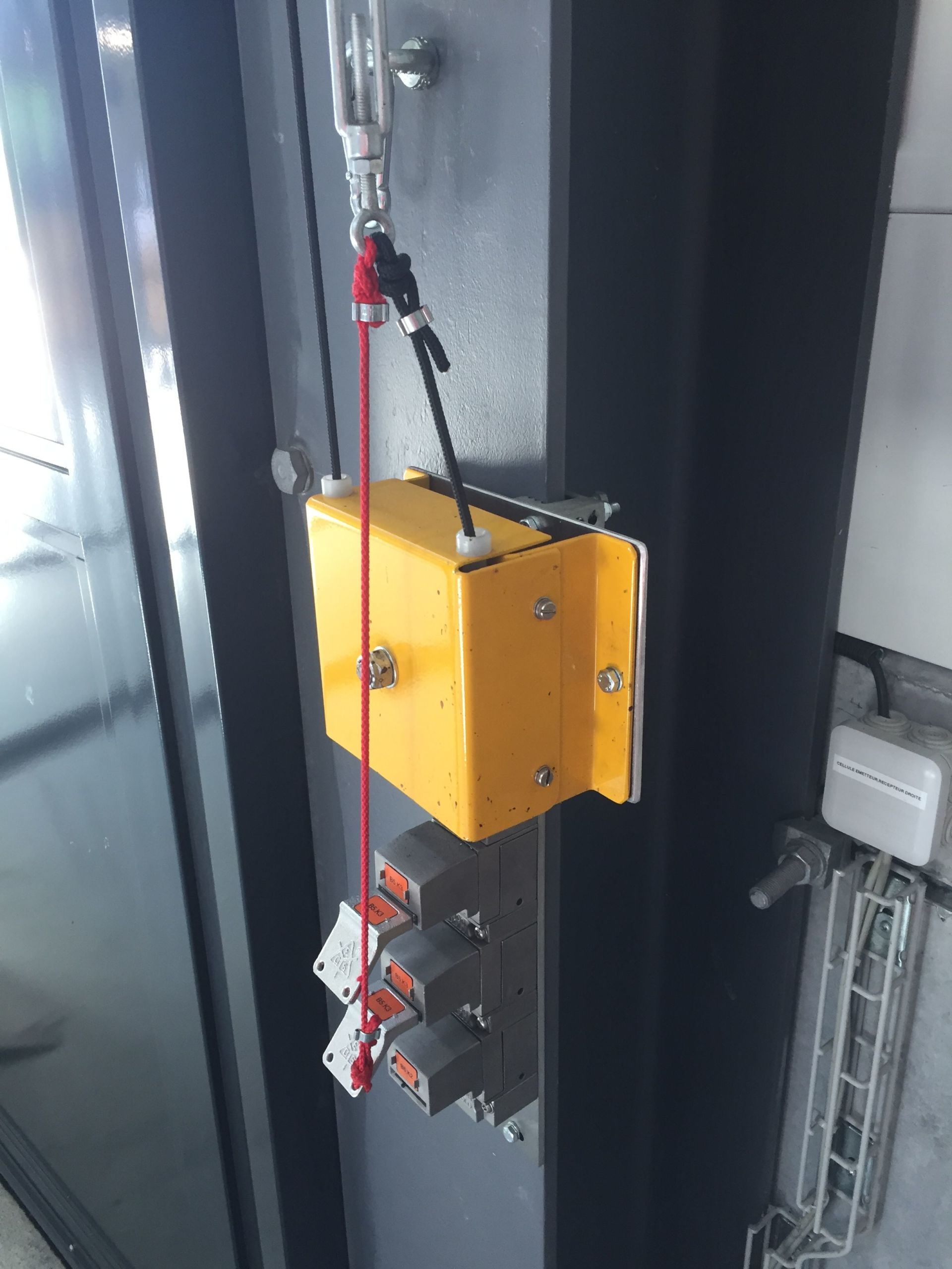 Gelber Sicherheitsschalter an einer Metallstruktur mit mehreren roten und silbernen Verriegelungsmechanismen und einer roten Zugschnur. Er scheint Teil eines industriellen oder mechanischen Systems zu sein.