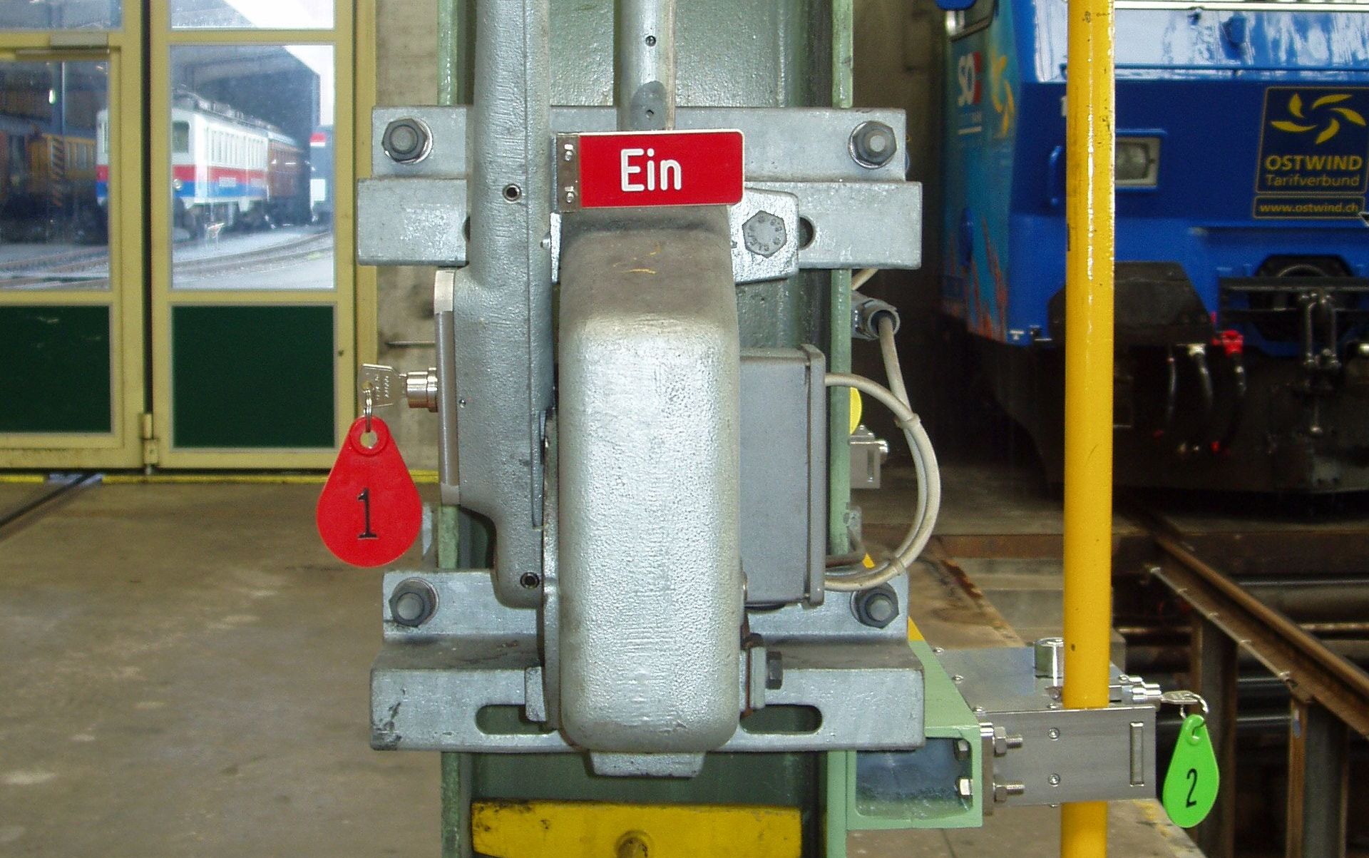 Eine metallene Maschinensteuerung in einem Bahnbetriebswerk, an der ein rotes Schild mit der Aufschrift „Ein“ und ein rotes Schild mit der Aufschrift „1“ befestigt sind. Im Hintergrund sind Gleise und mehrere Züge zu sehen, einer davon mit dem Logo „OSTWIND Tarifverbund“ und der Zahl „2“ auf einem grünen Schild am Gerät.