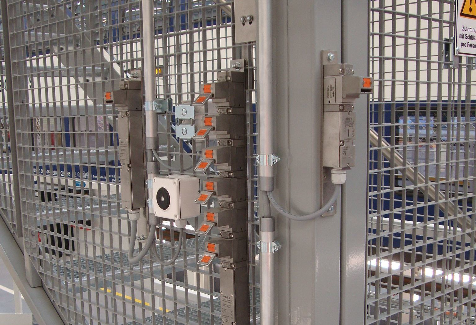 Eine Nahaufnahme eines Metallsicherheitstors mit Verriegelungsschaltern, Verkabelung und Bedienfeld. Der Aufbau umfasst graue und orangefarbene Komponenten, die auf einem vertikalen Metallrahmen innerhalb eines umzäunten Bereichs montiert sind. Oben im Bild ist teilweise ein Warnschild sichtbar.