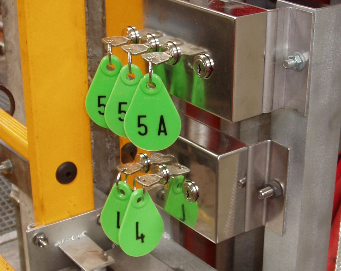 Mehrere grüne Schlüsselanhänger, jeder mit einer eindeutigen Nummer (z. B. 5, 5A, 14), hängen an glänzenden Metallhaken, die an einer metallischen Oberfläche befestigt sind. Der Hintergrund enthält eine gelbe vertikale Stützstruktur.