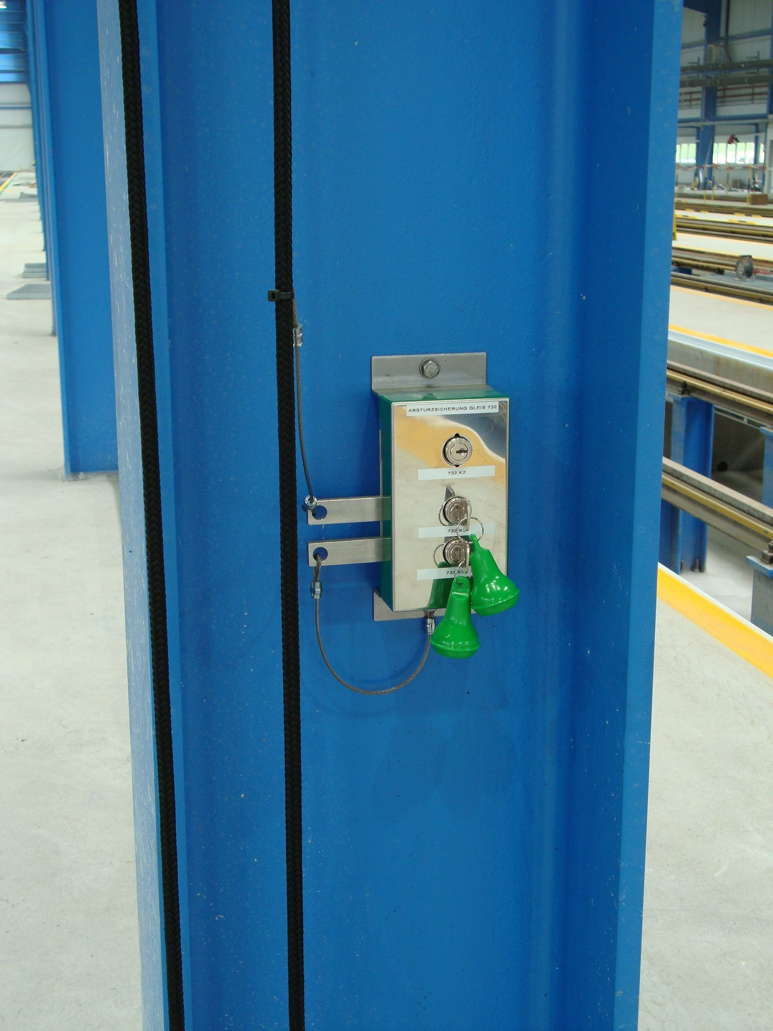 Ein blauer vertikaler Balken mit einem angebrachten industriellen Not-Aus-Tastensystem. Das System besteht aus einer Metallplatte mit zwei grünen Zuggriffen und roten Not-Aus-Tasten, montiert in einer industriellen Umgebung mit sichtbaren Schienen und Balken.