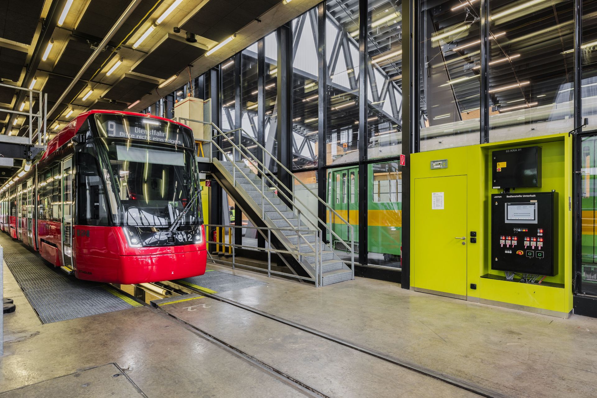 Eine moderne rote Straßenbahn steht in einer Wartungshalle. Daneben befinden sich eine Metalltreppe und ein Gehweg, die zu einer oberen Etage führen. Die Halle hat Glaswände, große Fenster und verschiedene Bedienfelder. Im Hintergrund ist eine weitere grüne Straßenbahn zu sehen.