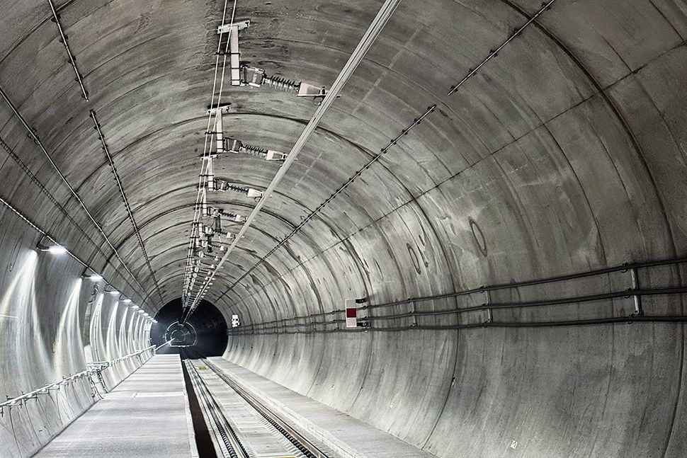 Ein Blick in einen grossen, leeren Eisenbahntunnel mit Eisenbahnschienen, Deckenbeleuchtung, neuer Deckenstromschiene an der Tunneldecke und Kabeln, die an den geschwungenen Wänden entlangverlaufen und so ein Gefühl von Tiefe und Perspektive vermitteln.