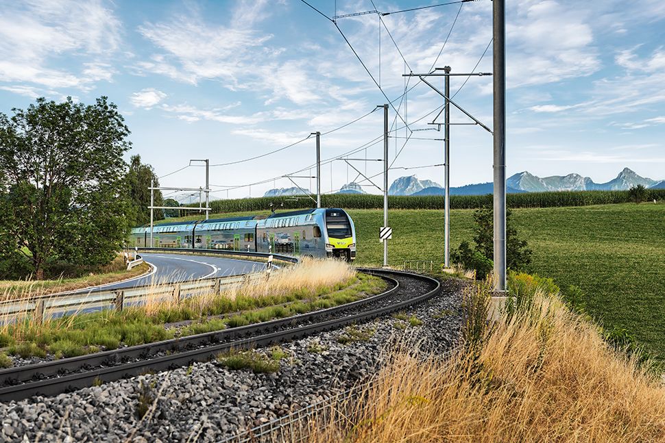 Ein moderner Personenzug fährt auf kurvigen Gleisen durch grüne Felder, im Hintergrund sind Berge zu sehen, und über ihm erstreckt sich ein teilweise bewölkter Himmel.