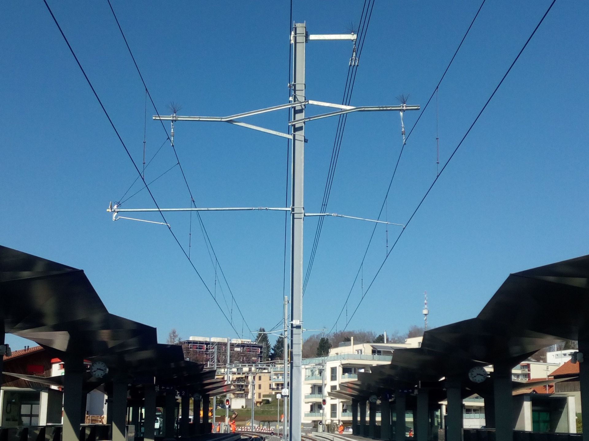 Eine symmetrische Ansicht eines Bahnhofs mit Oberleitung. Die Metallmasten und -leitungen bilden ein geometrisches Muster vor einem klaren blauen Himmel. Die Bahnsteige sind mit modernen Unterständen gesäumt und im Hintergrund sind Gebäude zu sehen.
