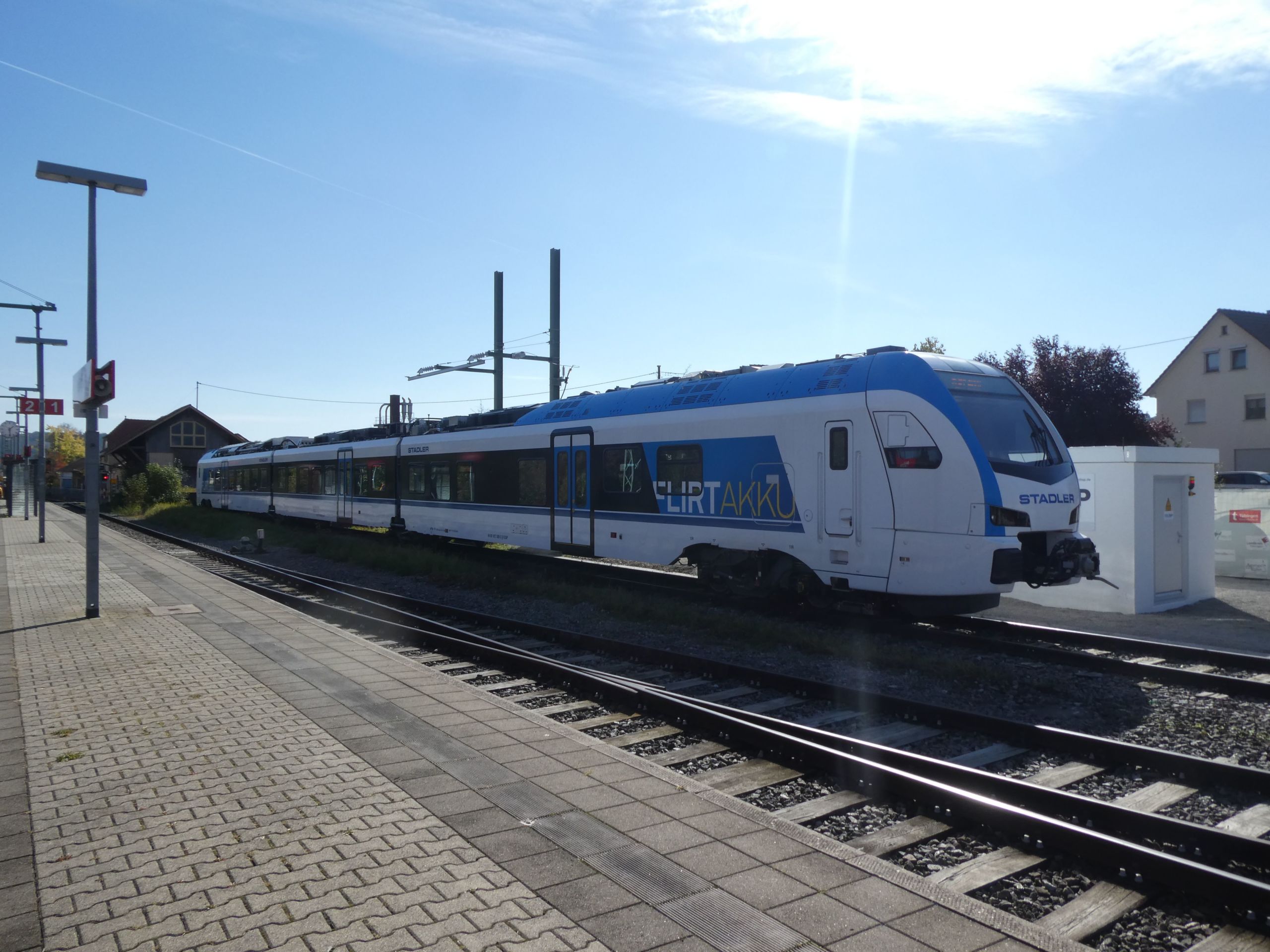 Ein moderner blau-weißer Personenzug steht an einem sonnigen Bahnhof mit klarem blauen Himmel über ihm. Der Bahnsteig ist leer, im Hintergrund sind ein paar Häuser zu sehen. Auf der Seite des Zuges steht das Wort „ILIRTATIR“.