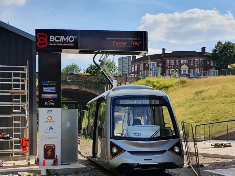 Eine moderne Straßenbahn auf einer Testanlage mit der Aufschrift BCIMO steht unter einem Hightech-Bogen mit Logos und Informationen. Im Hintergrund sind ein grasbewachsener Damm, eine gemauerte Eisenbahnbrücke und ein altes Gebäude mit der Aufschrift „STATION HOTEL“ zu sehen. Klarer Himmel mit einigen Wolken darüber.